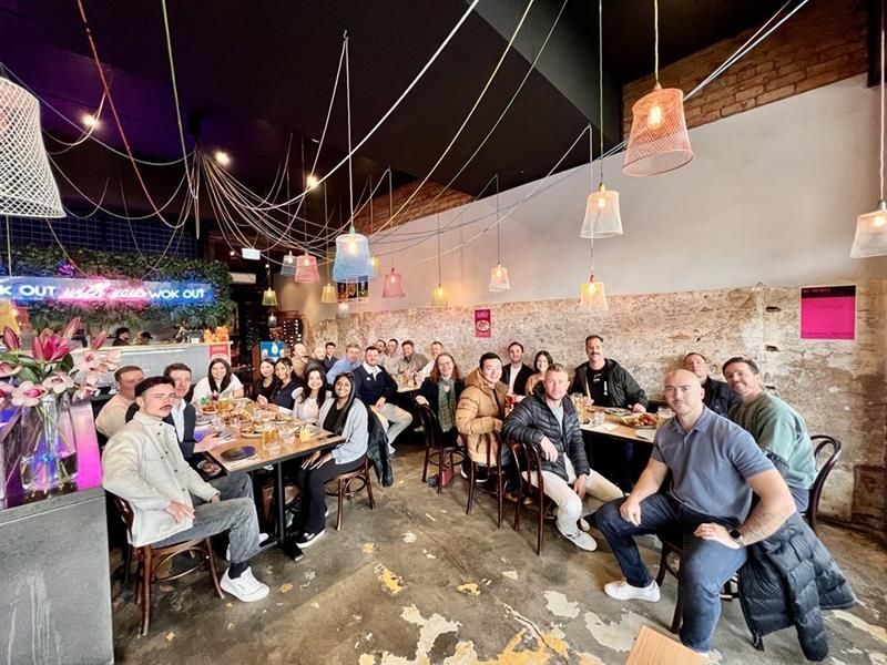 Group of people at a restaurant, sitting at tables. Some are smiling. The interior is decorated with lights.