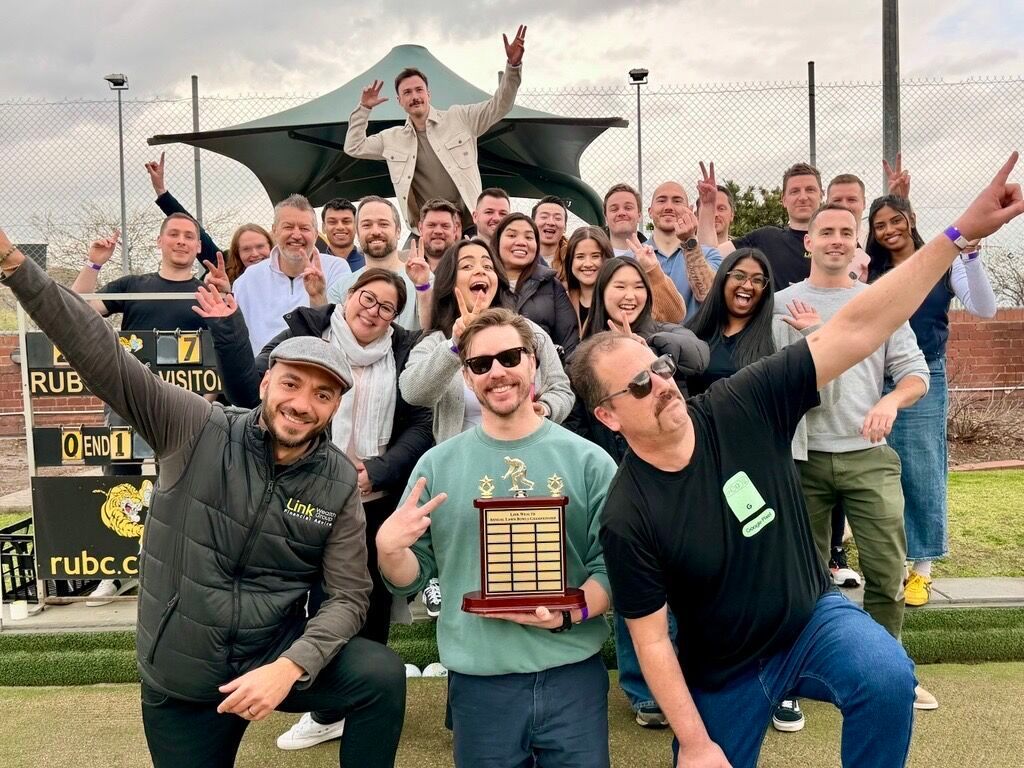 Group celebrates a win outdoors, holding a trophy. People smiling, raising arms. Green grass and a black canopy.