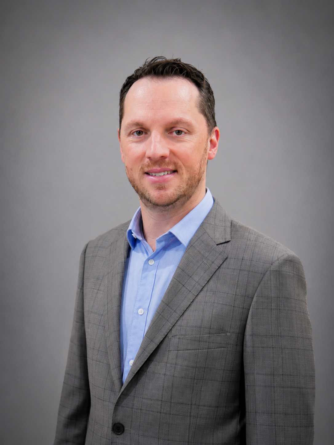 Man in a gray suit and blue shirt smiles, standing against a wood-paneled wall.