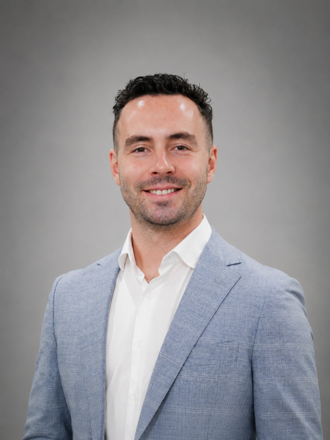 Man in blue suit and white shirt poses with hands in pockets, against a wood panel backdrop.