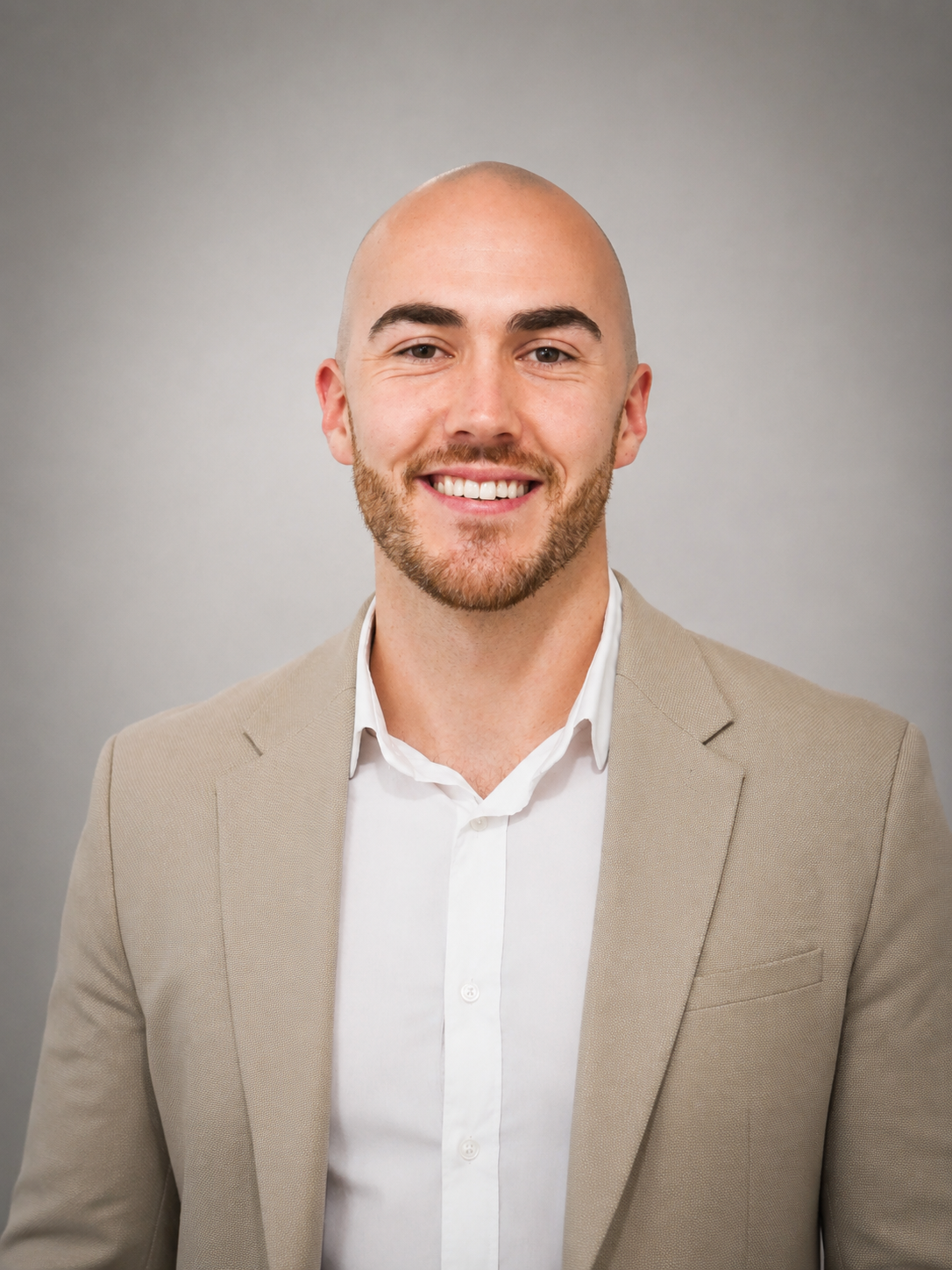 Bald man in beige blazer, white shirt, and dark pants poses with hands in pockets against a wood panel background.