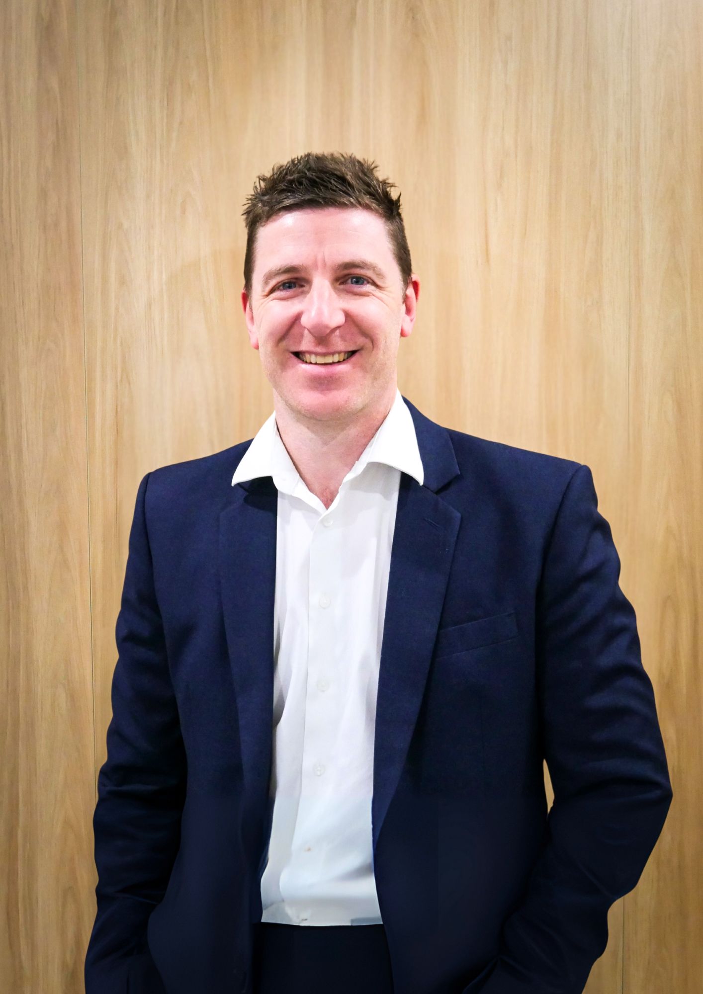 Man in a navy suit and white shirt smiles in front of a wood-paneled wall.