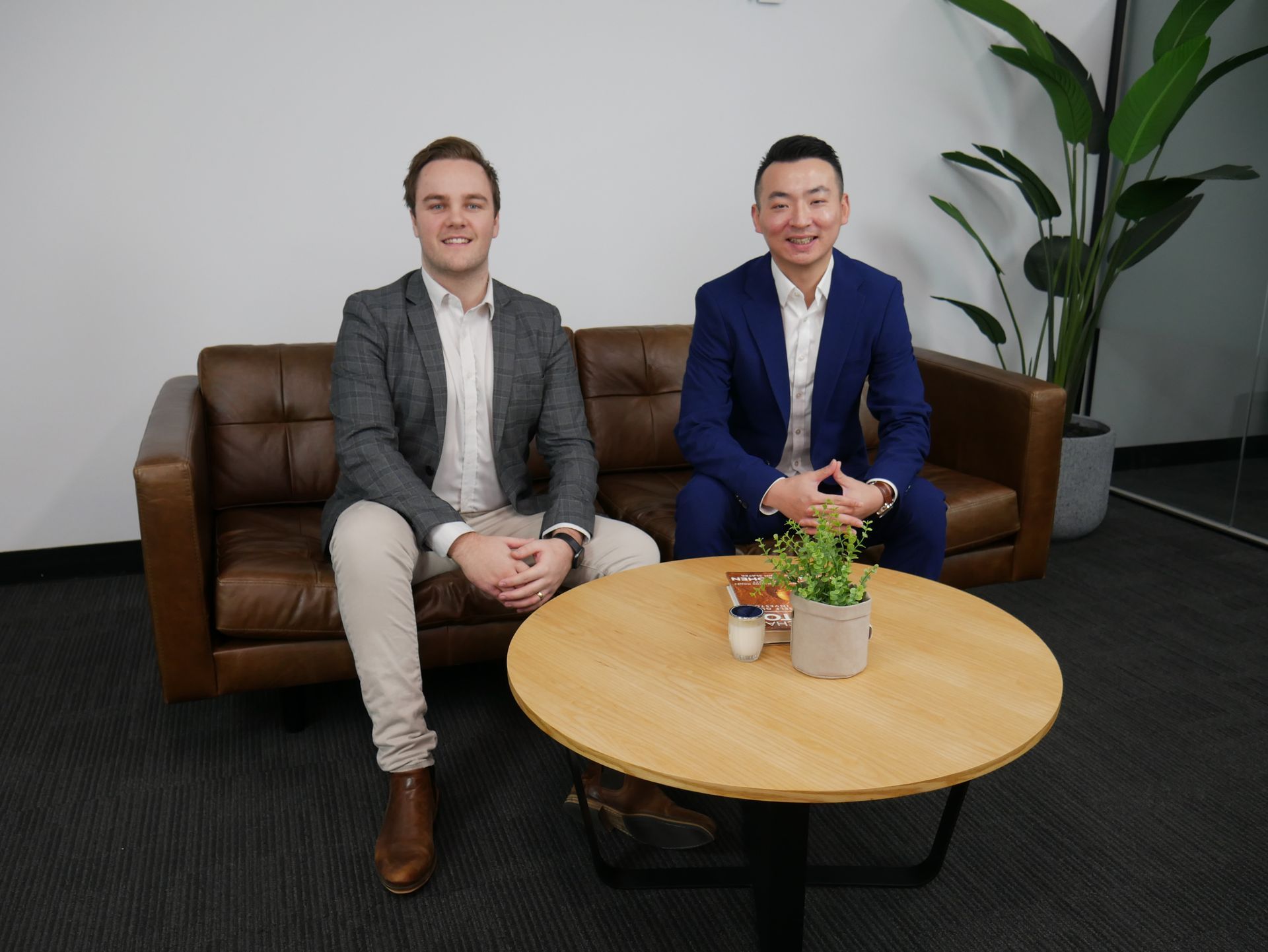Two men in business attire sit on a brown sofa near a round table with plants, indoors.
