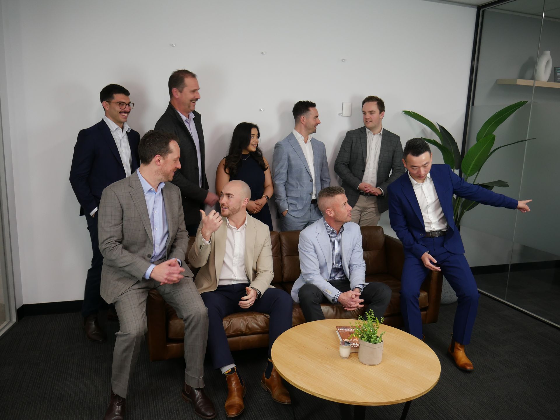 Group of nine people in suits, posing in an office. Some are standing, others sitting on a couch, with a table in front.