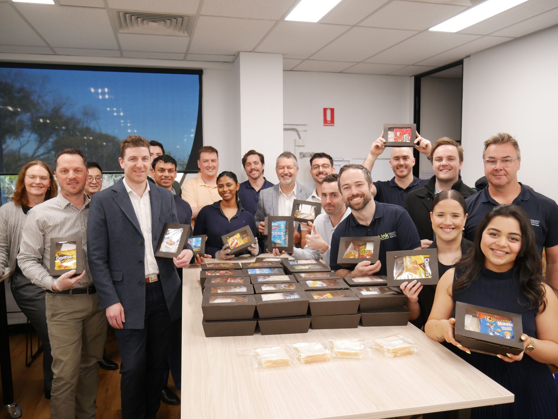 Group of people in an office holding boxes of treats, smiling. Boxes of food are on a table.