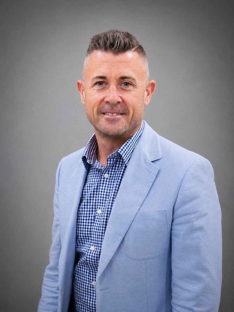 Man in light blue blazer and patterned shirt stands in front of a wood-paneled wall.