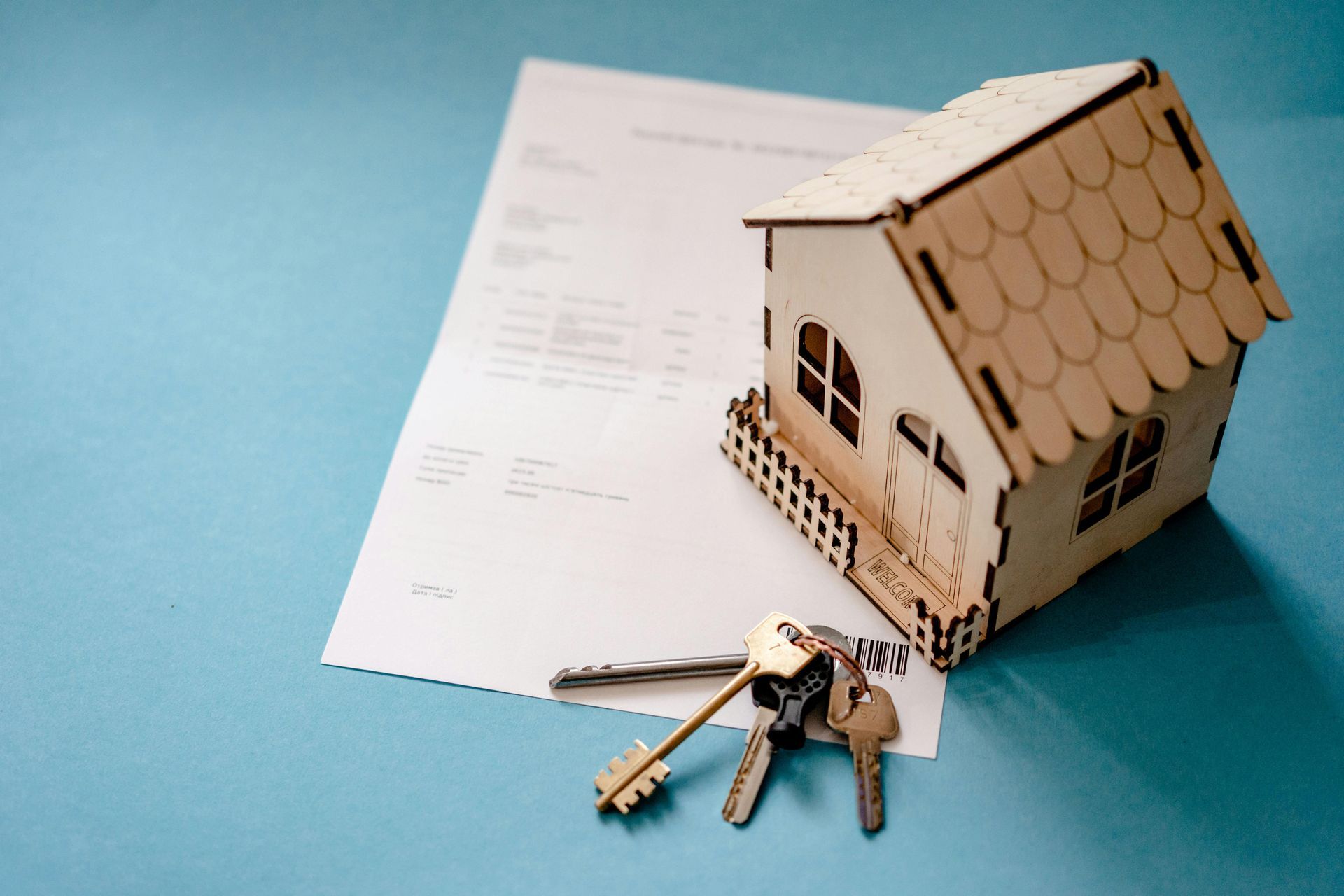 A wooden house model, keys, and a document on a blue surface, symbolizing a real estate transaction.