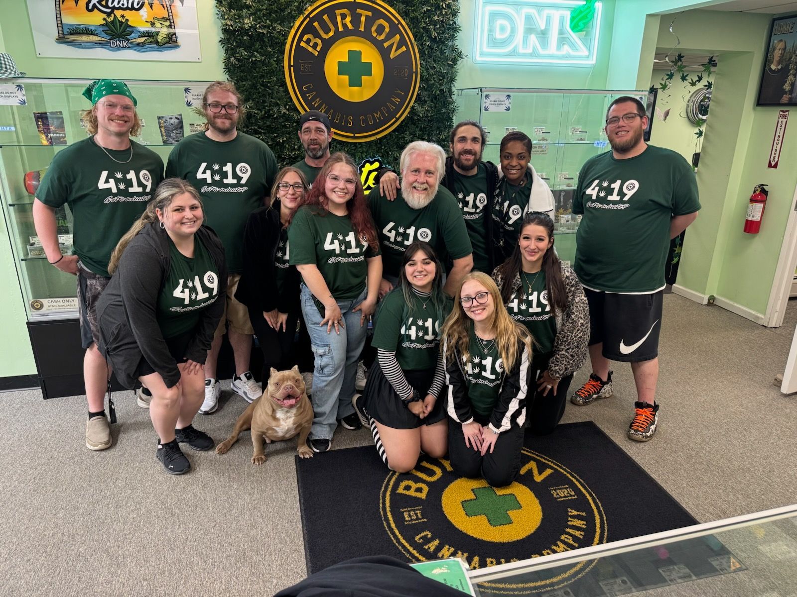 Group of people in green shirts pose in a cannabis shop; a dog sits on the floor near them.