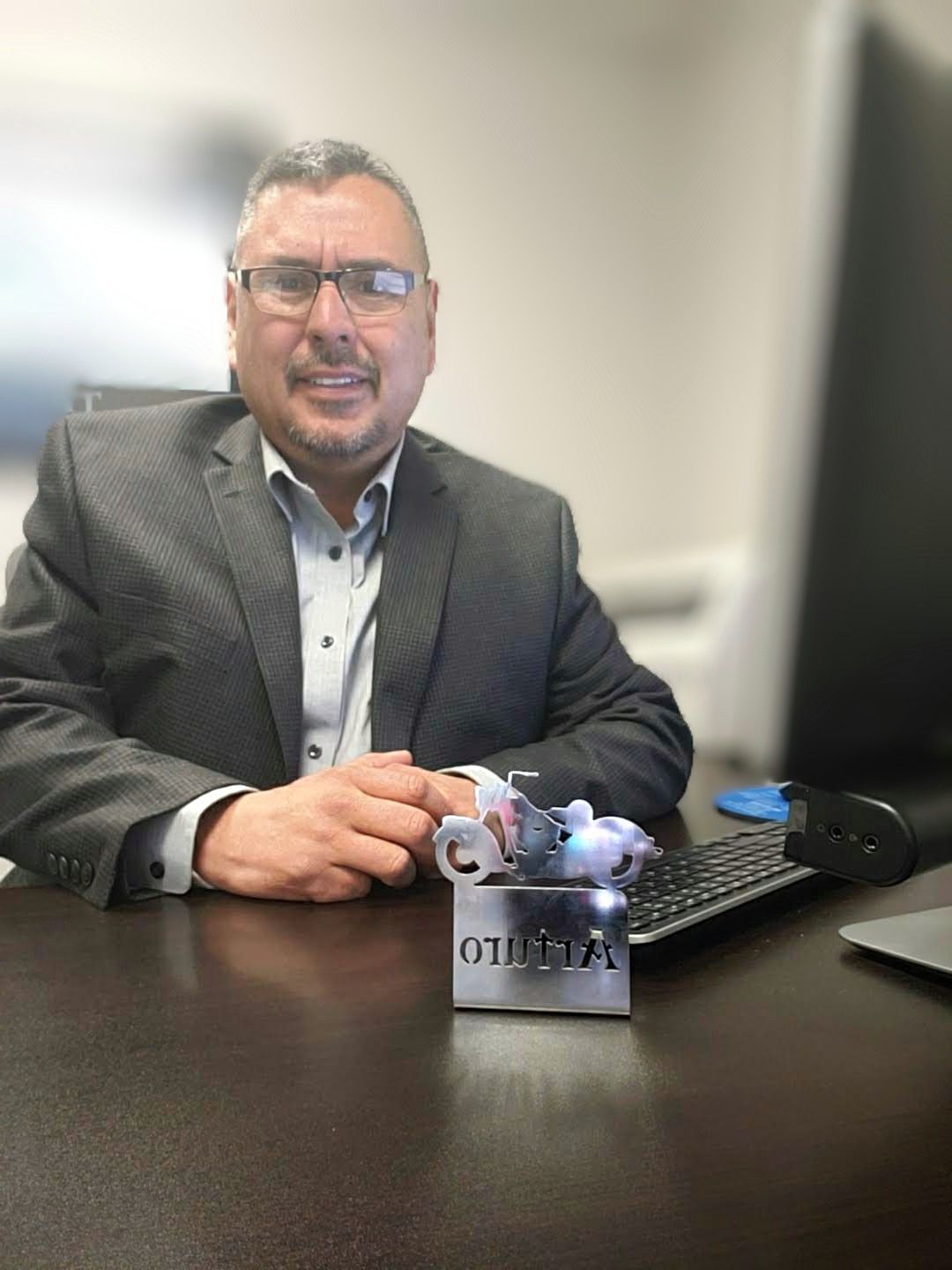 Man in a gray blazer seated at a desk with a small robot figurine in an office setting