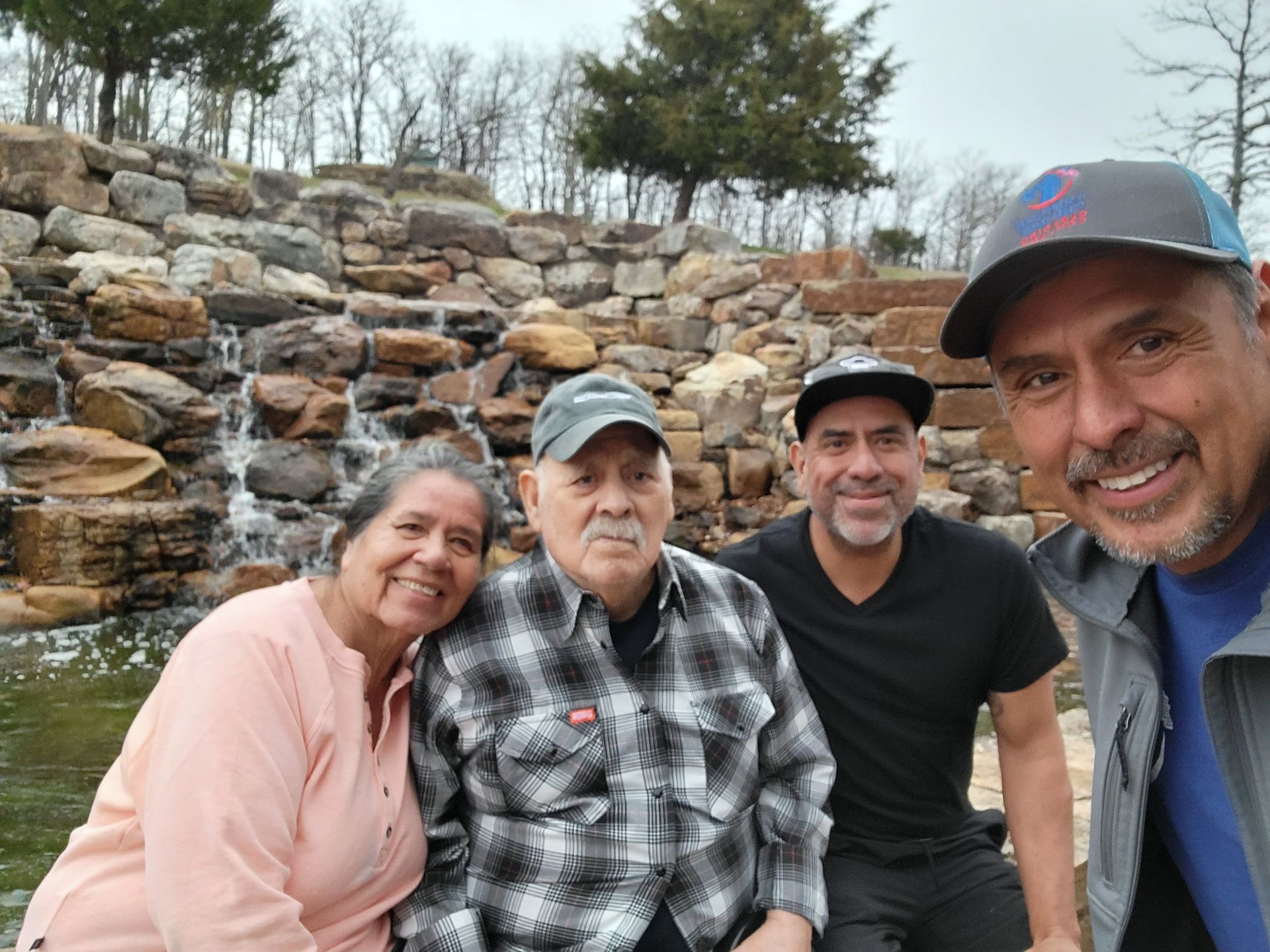 Four people smiling outdoors by a stone waterfall, with trees and water in the background.
