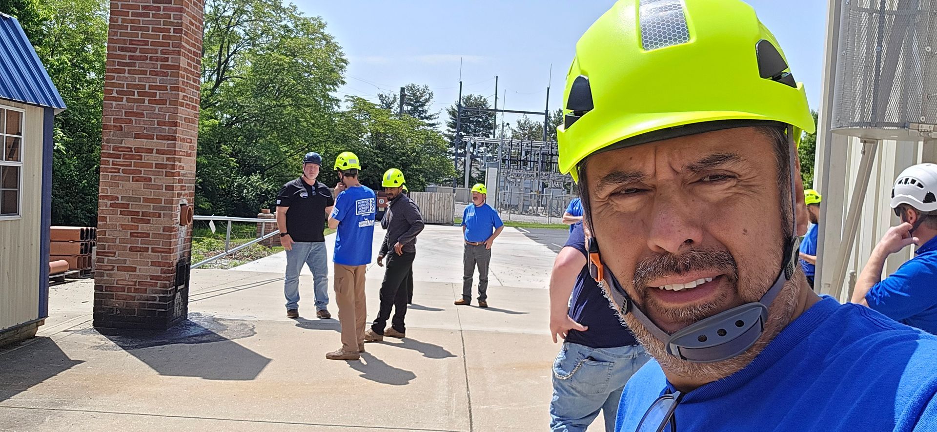 Construction worker in neon hard hat taking a selfie at a building site with several people behind him