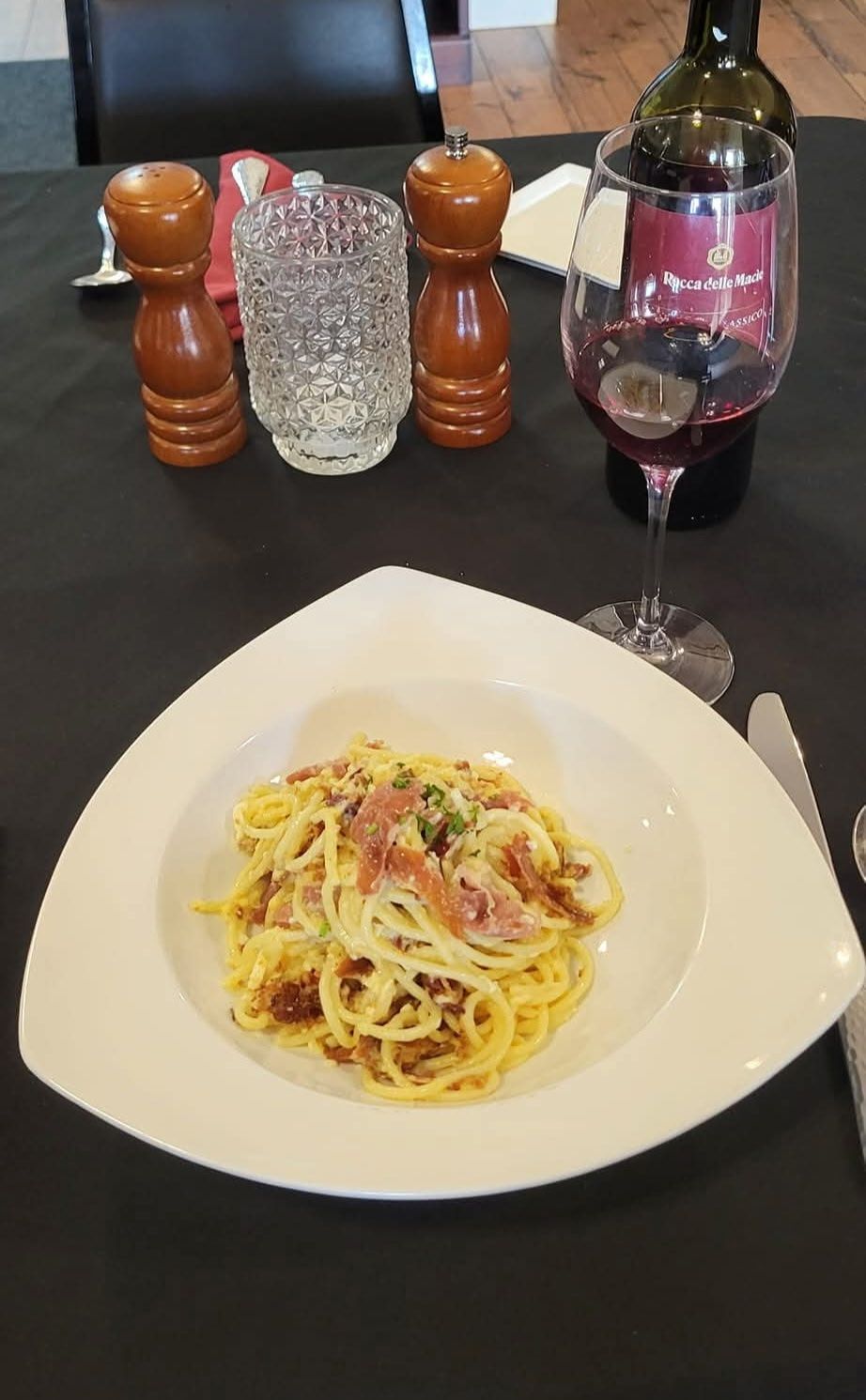 Pasta carbonara in a white triangular bowl, with wine and condiments on a black tablecloth.