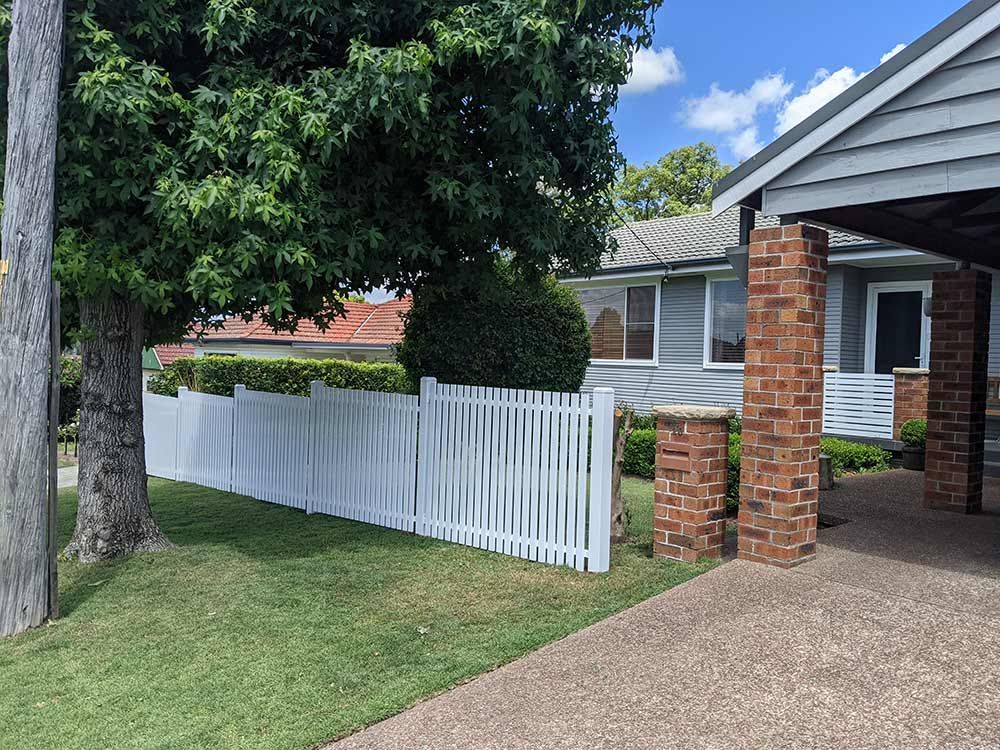 A White Picket Fence is in Front of a Brick House — Rearden Fencing Pty Ltd in Toronto, NSW
