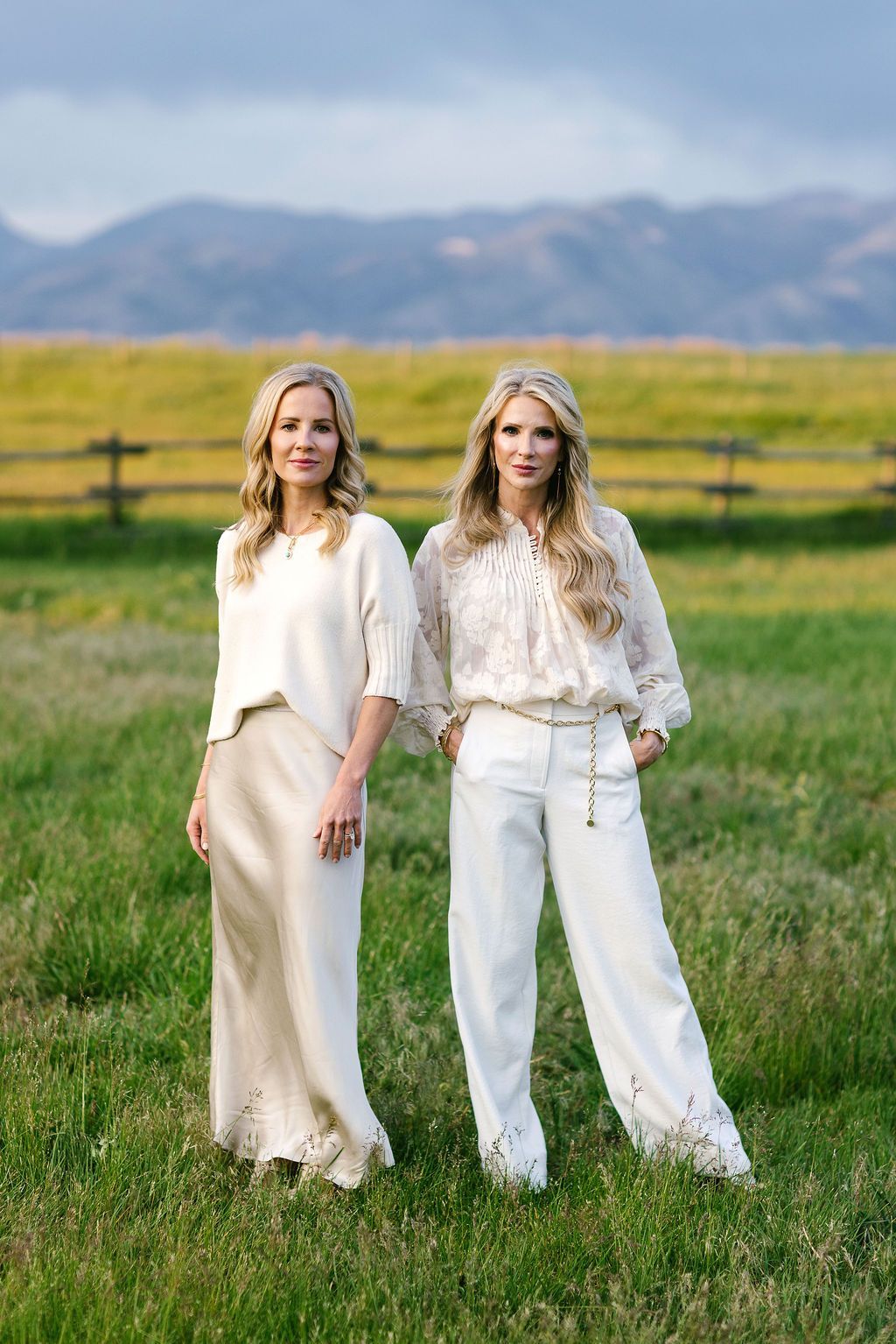 Two women in cream outfits standing in a grassy field with mountains in the background