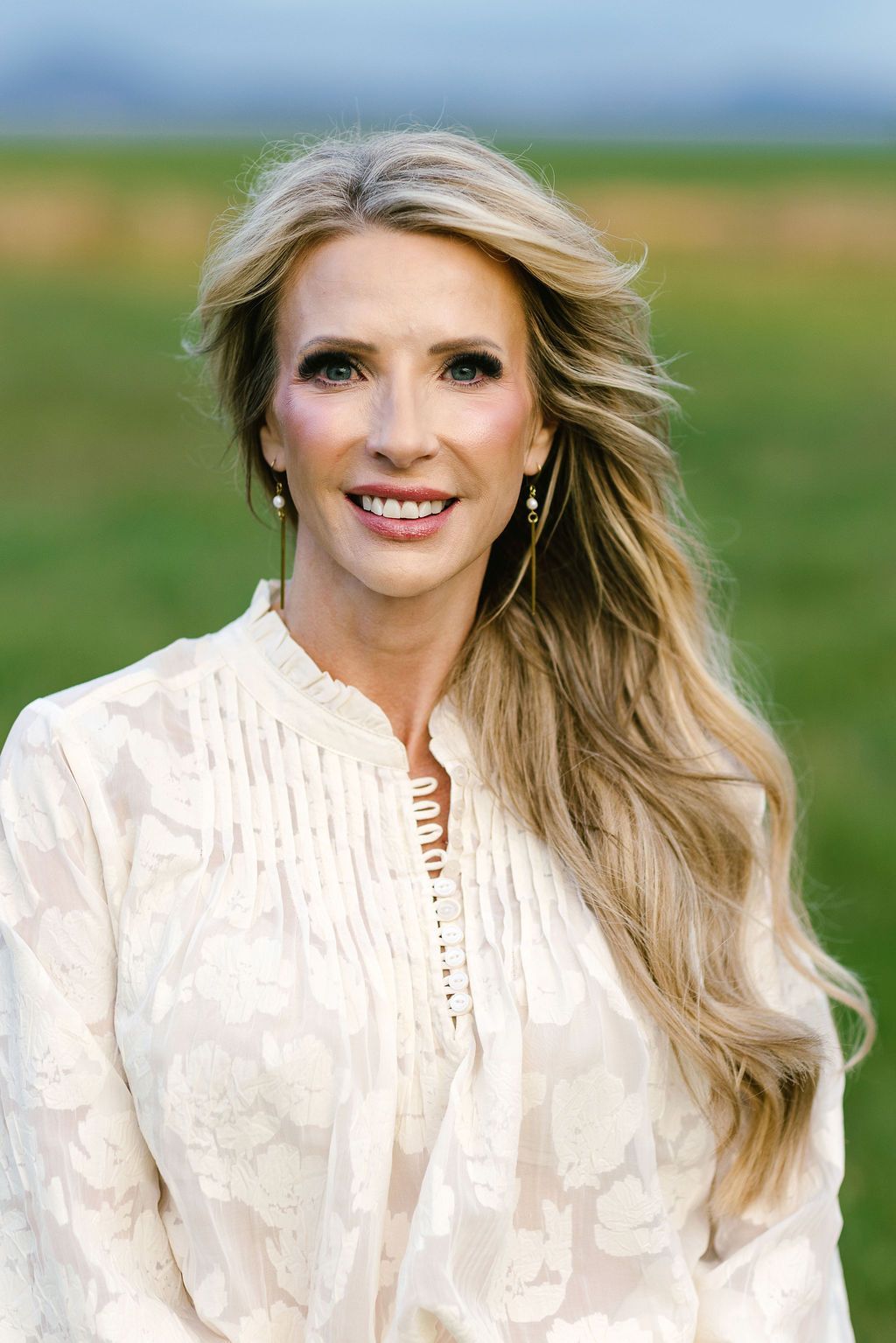 Portrait of a smiling woman in a white blouse, standing outdoors with a blurred field and blue sky behind her