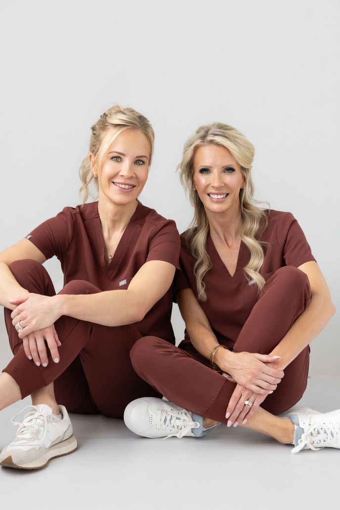 Two women in matching green outfits sit on white chairs in a stylish hallway with artwork and a doorway.