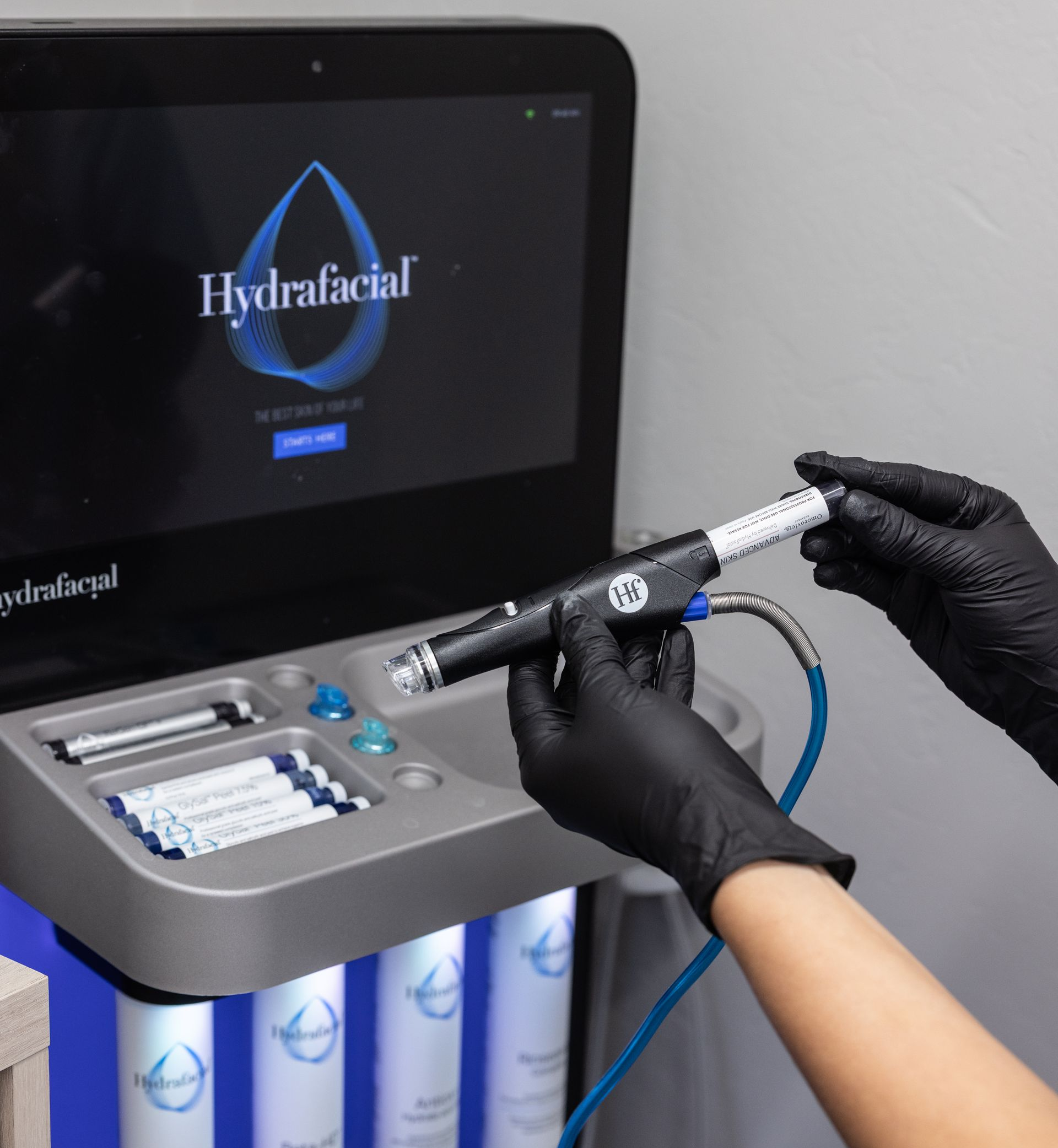 Gloved hands hold a dental handpiece beside a HydraFacial machine with treatment tools and tubing.