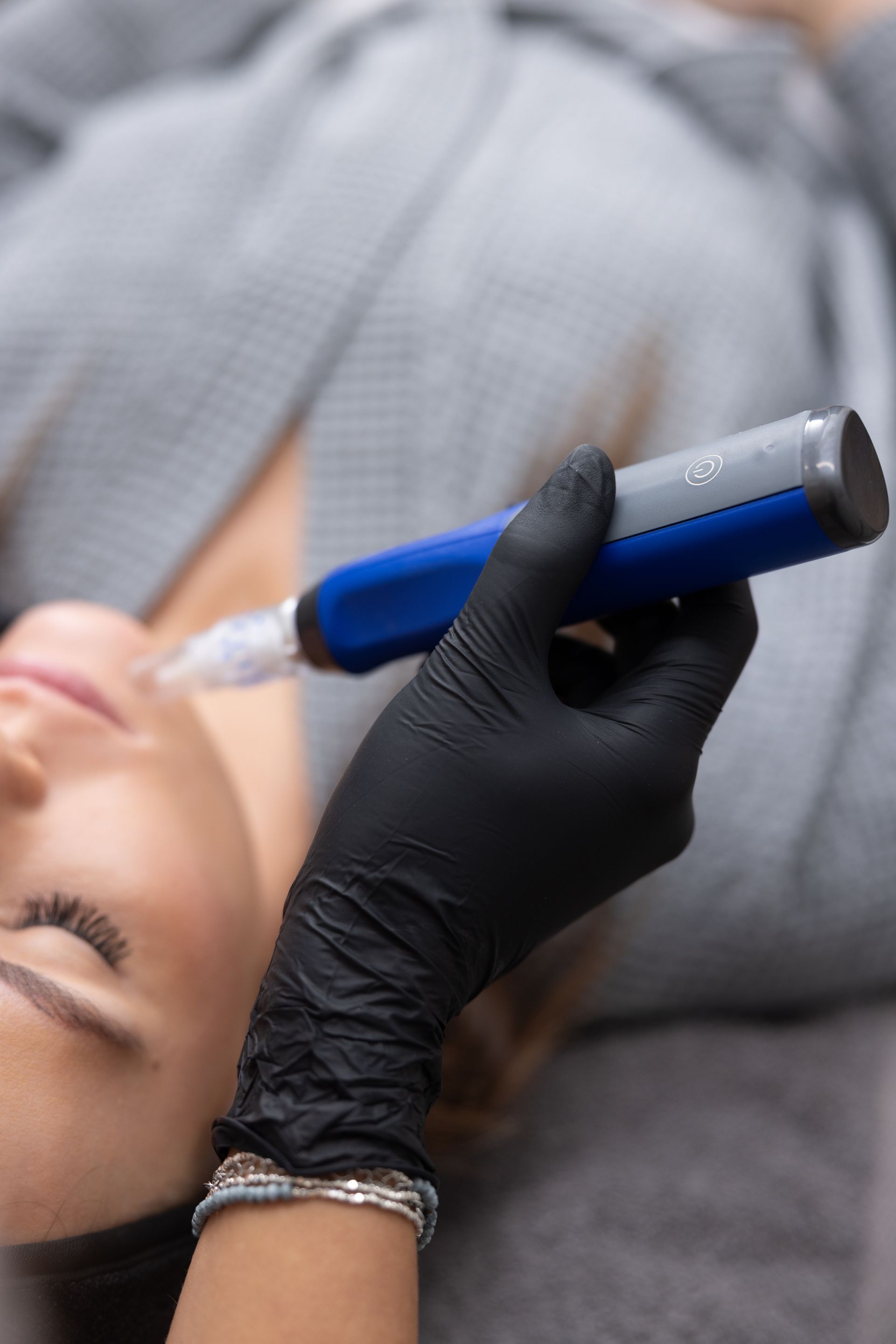A practitioner in black gloves uses a blue dermaplaning tool on a reclining client's face.