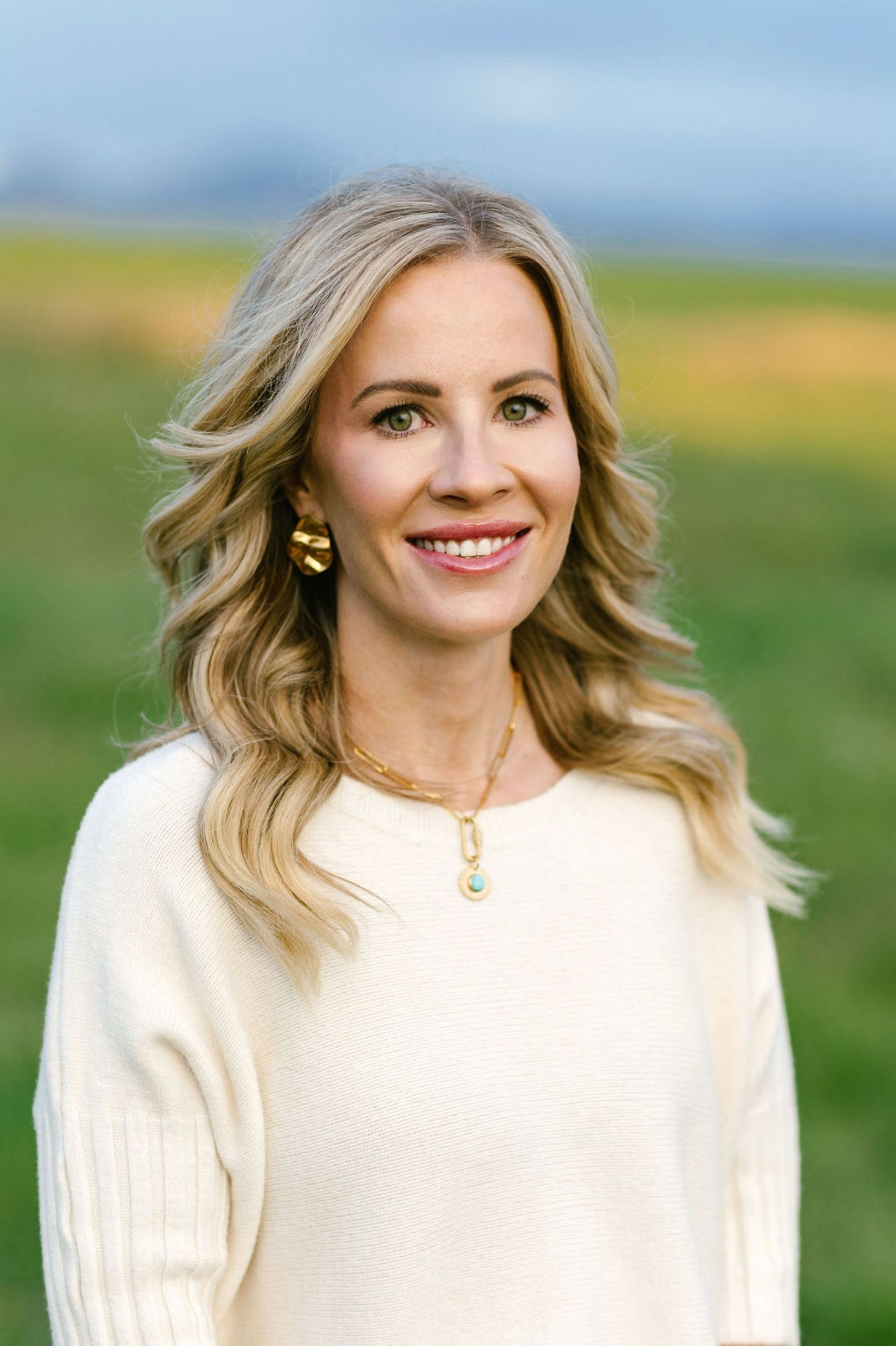 Smiling woman in a cream sweater standing outdoors in a grassy field with a blurred horizon