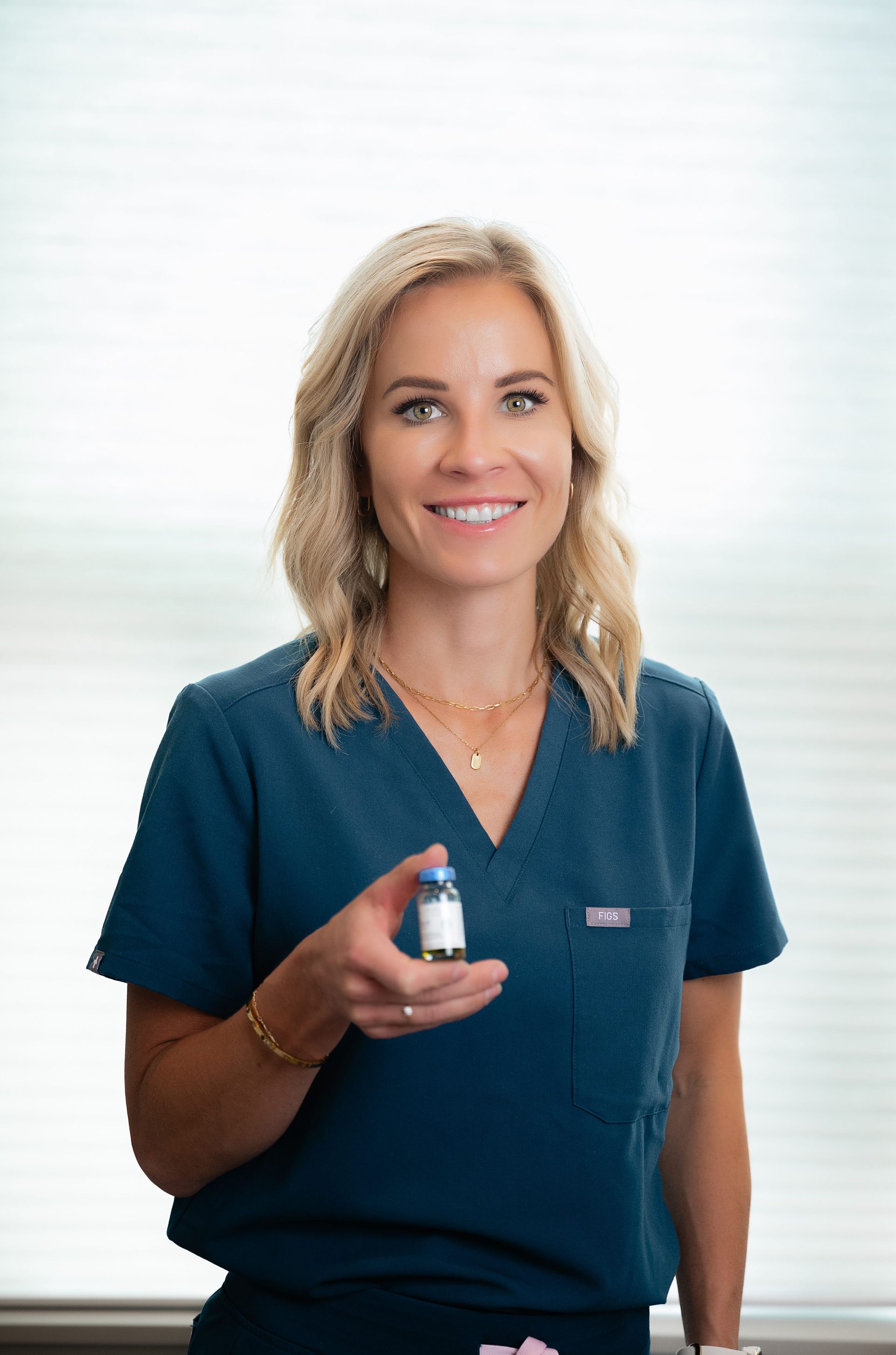 Woman in teal scrubs holding a small vial, smiling against a bright white background.