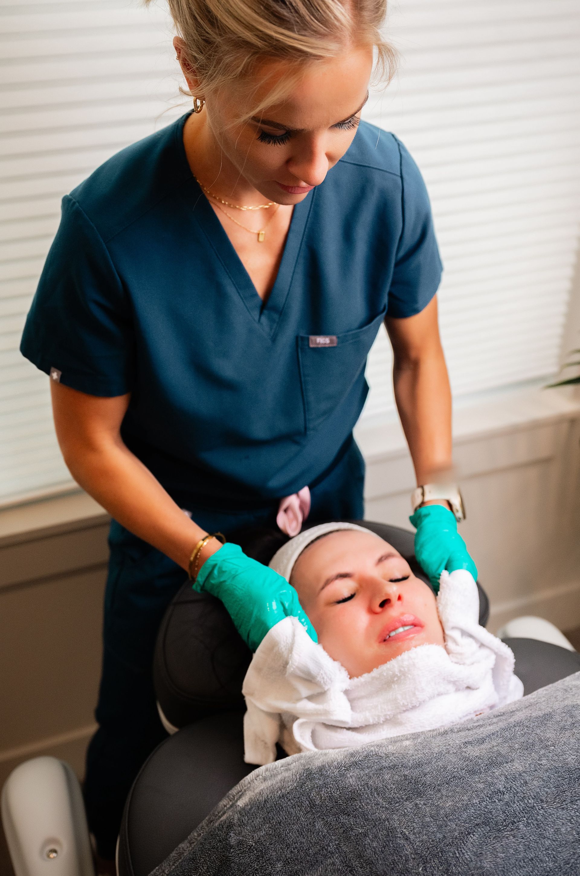 A clinician in teal scrubs cleans a patient’s face with gloved hands in a treatment room.