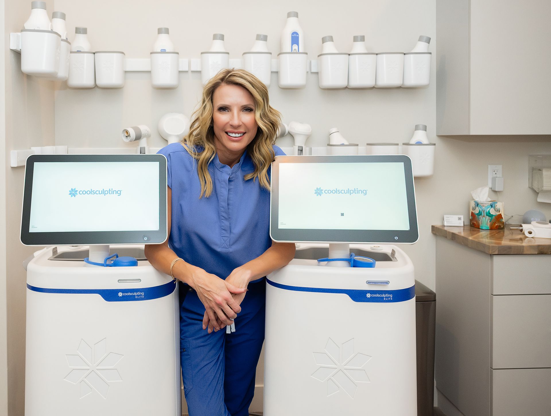 Woman in blue scrubs smiling between two medical machines in a clinic room
