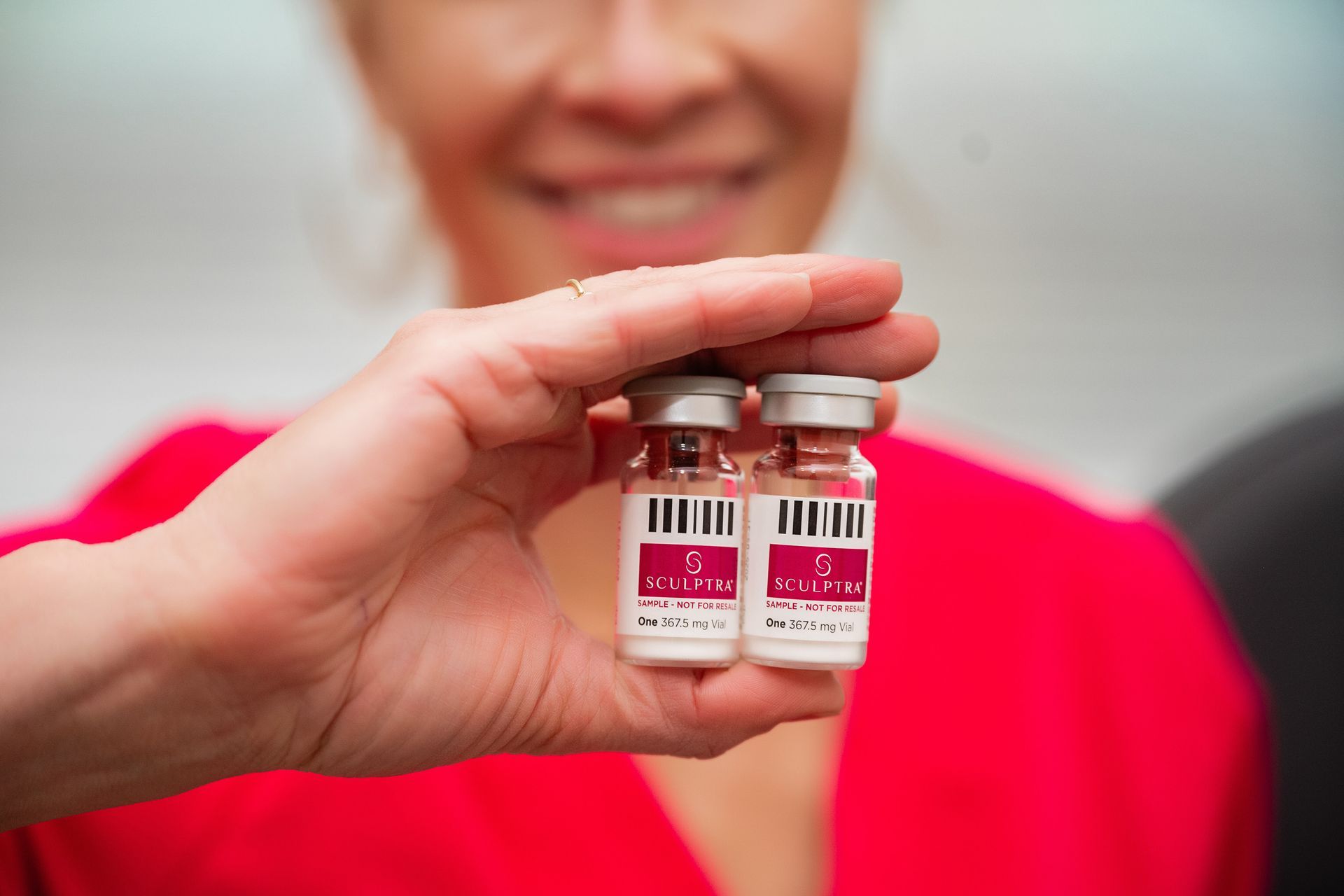Hand holding two small vaccine vials in front of a smiling person in a red shirt