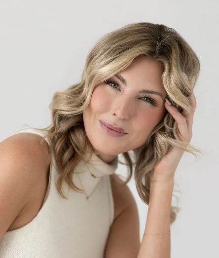 Portrait of a smiling woman in a cream top, resting her hand near her face against a light background