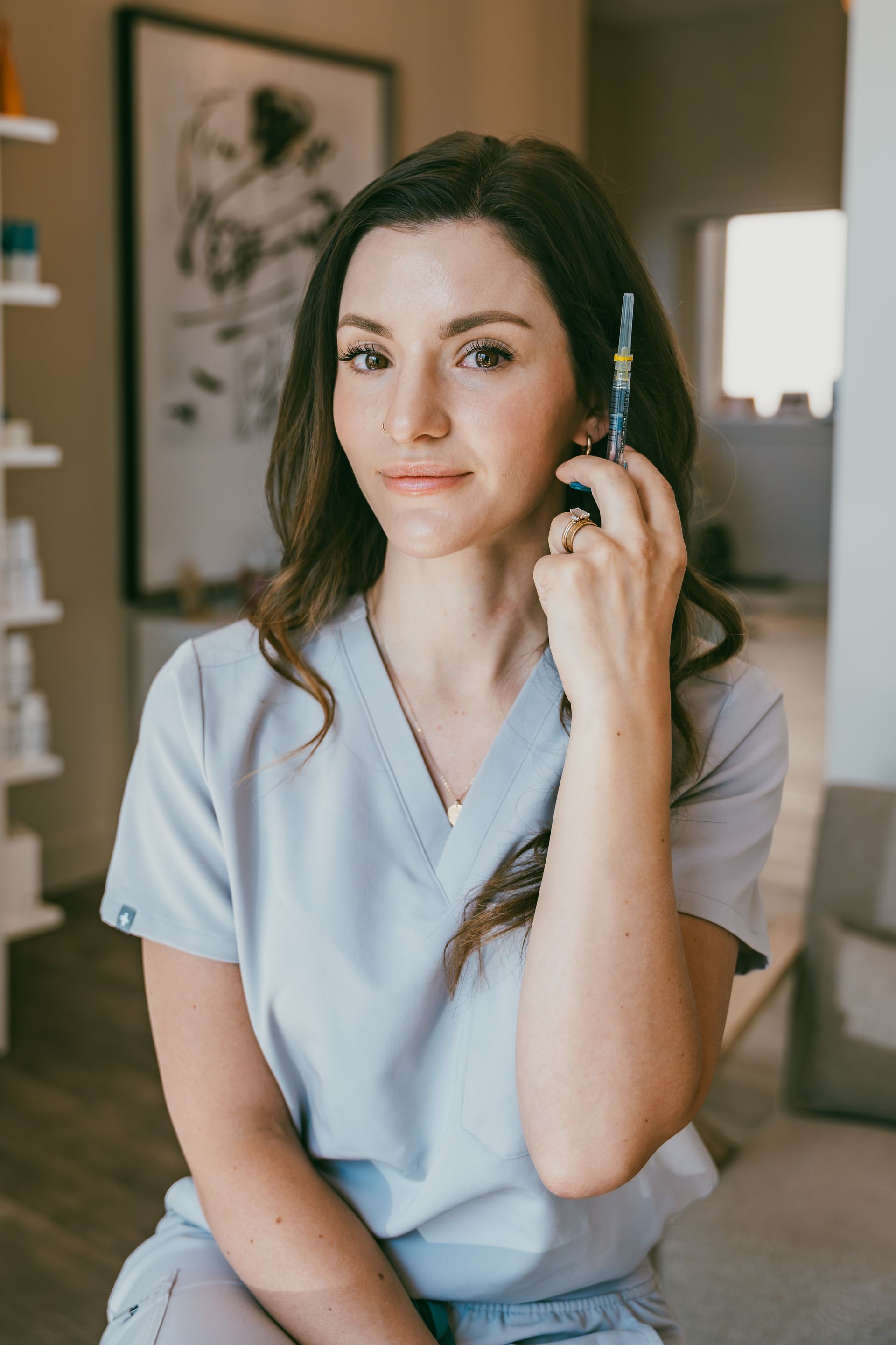Woman in light blue scrubs holding a syringe, seated indoors near a window with a calm expression