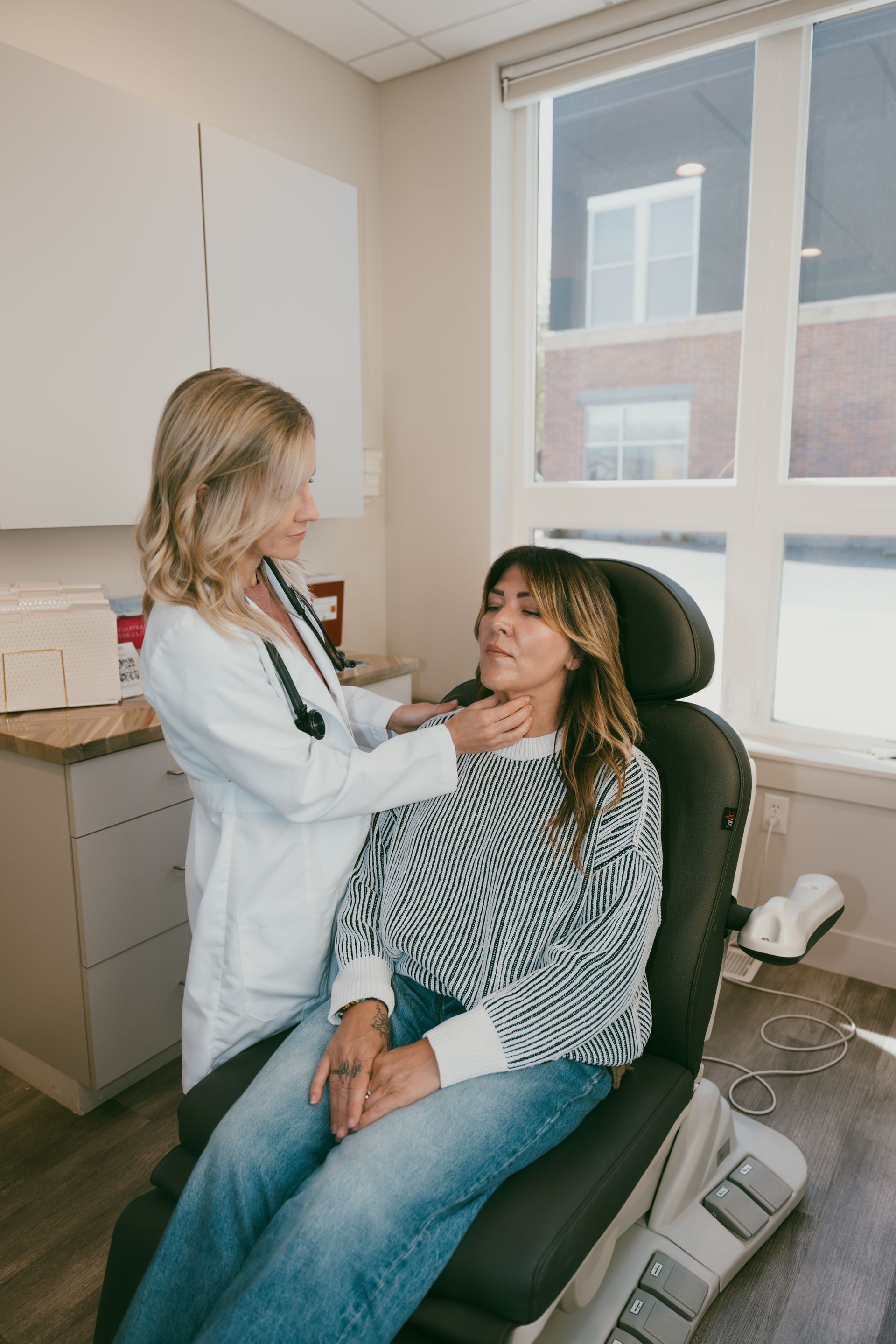 Doctor examines a seated patient in a bright exam room by a window.