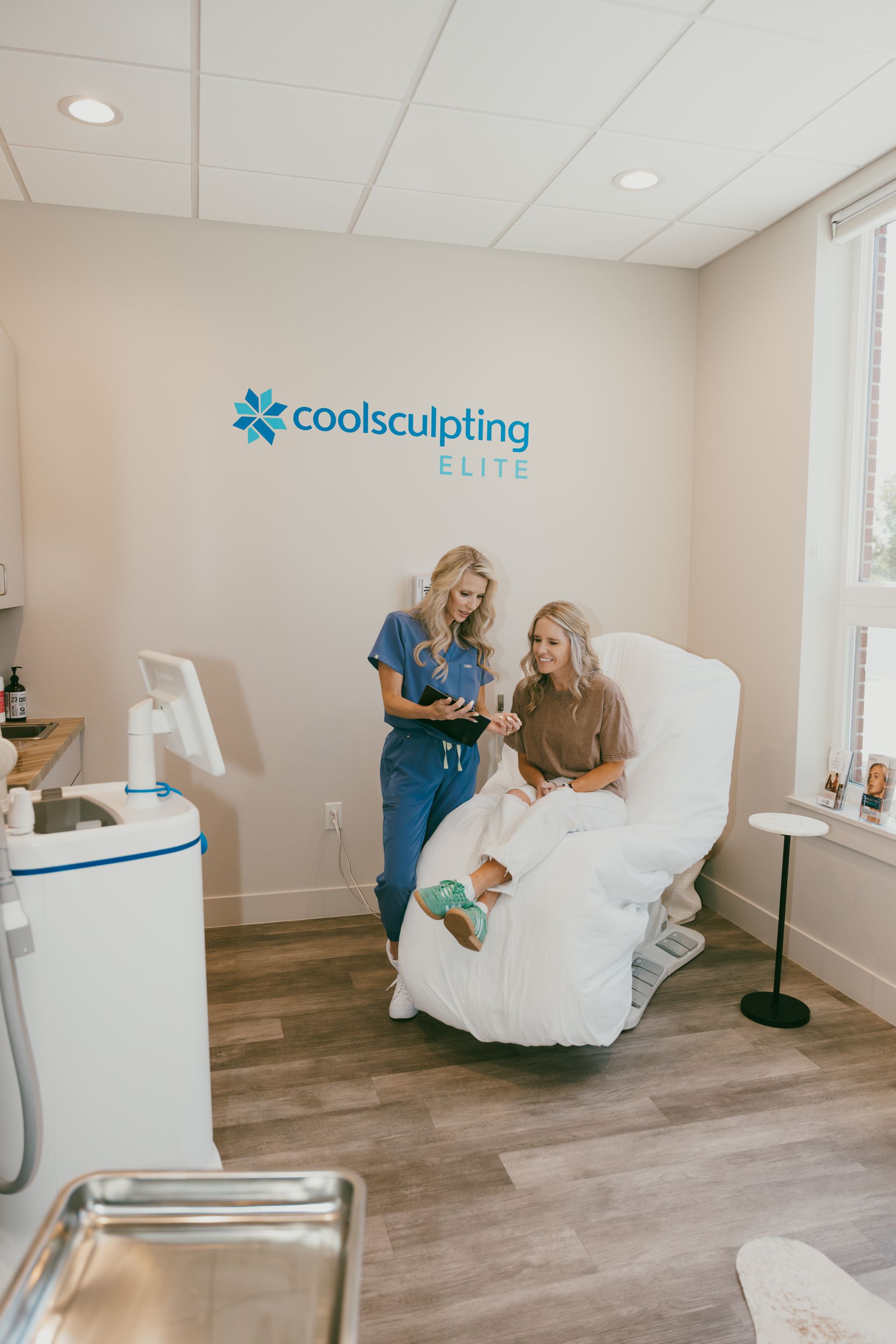 Two people in a CoolSculpting clinic, one seated on a treatment chair and one standing beside her.
