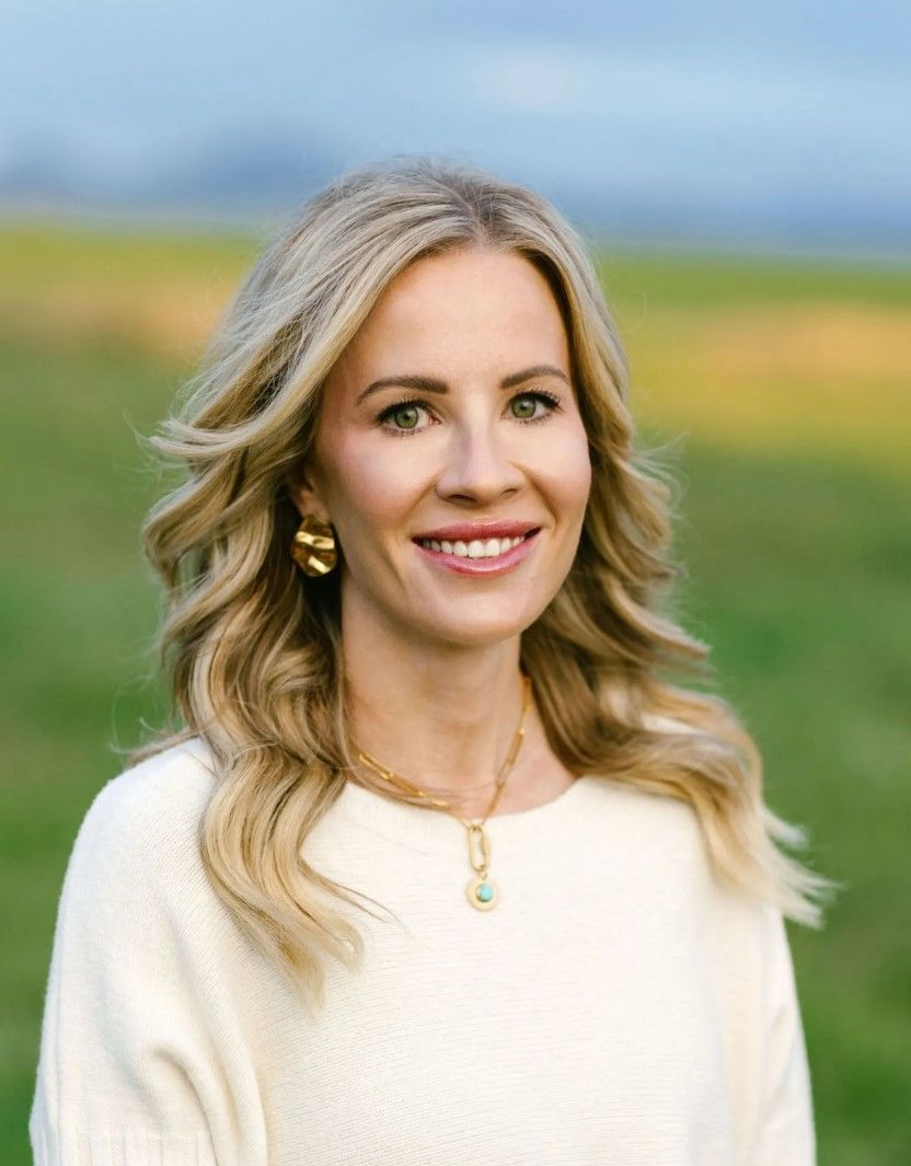 Smiling woman in a white sweater standing outdoors in a grassy field with a blurred mountain backdrop