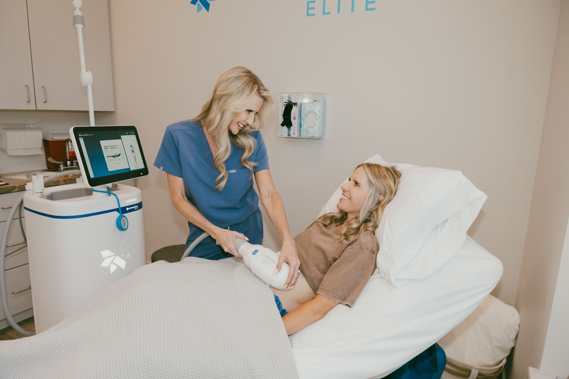 A clinician assists a reclining patient in a medical spa room with a treatment device nearby.