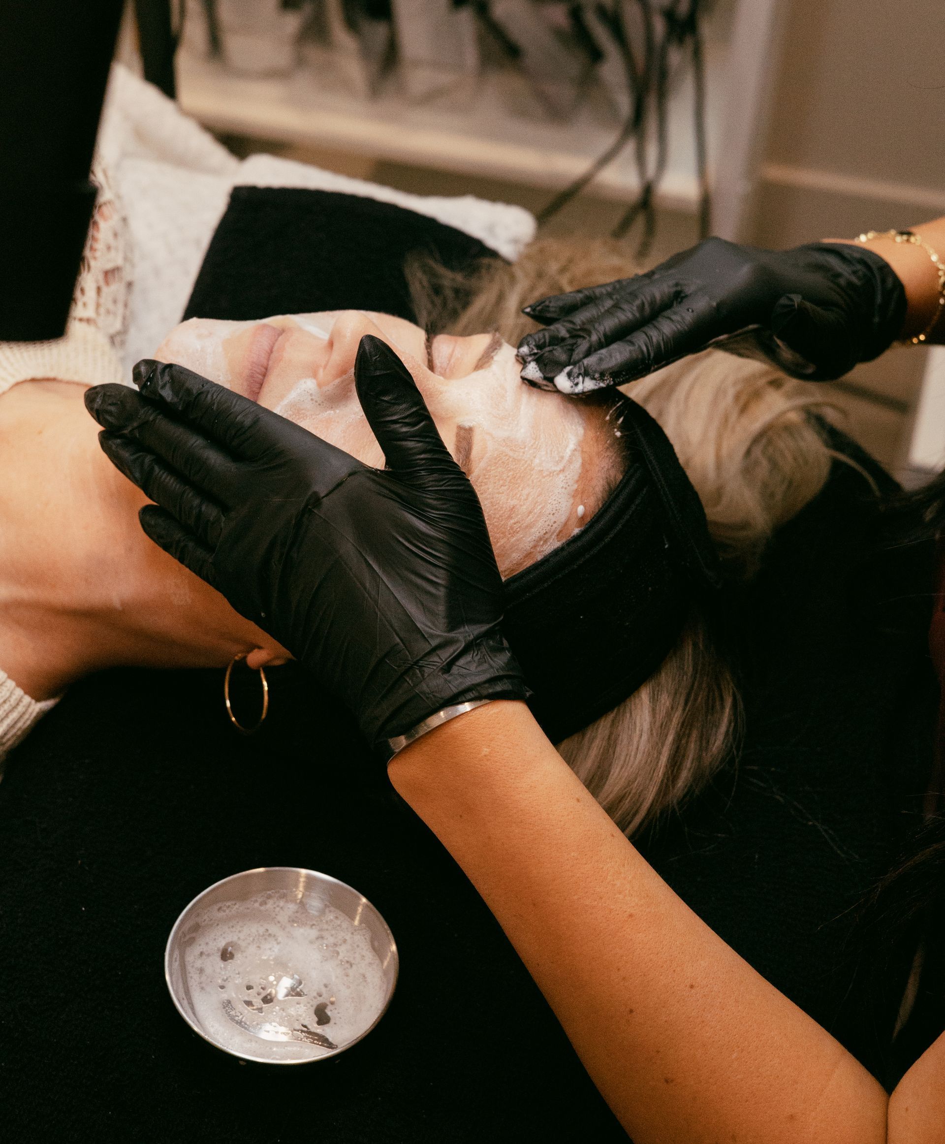 Gloved hands applying a facial treatment to a client lying back with a towel around the head