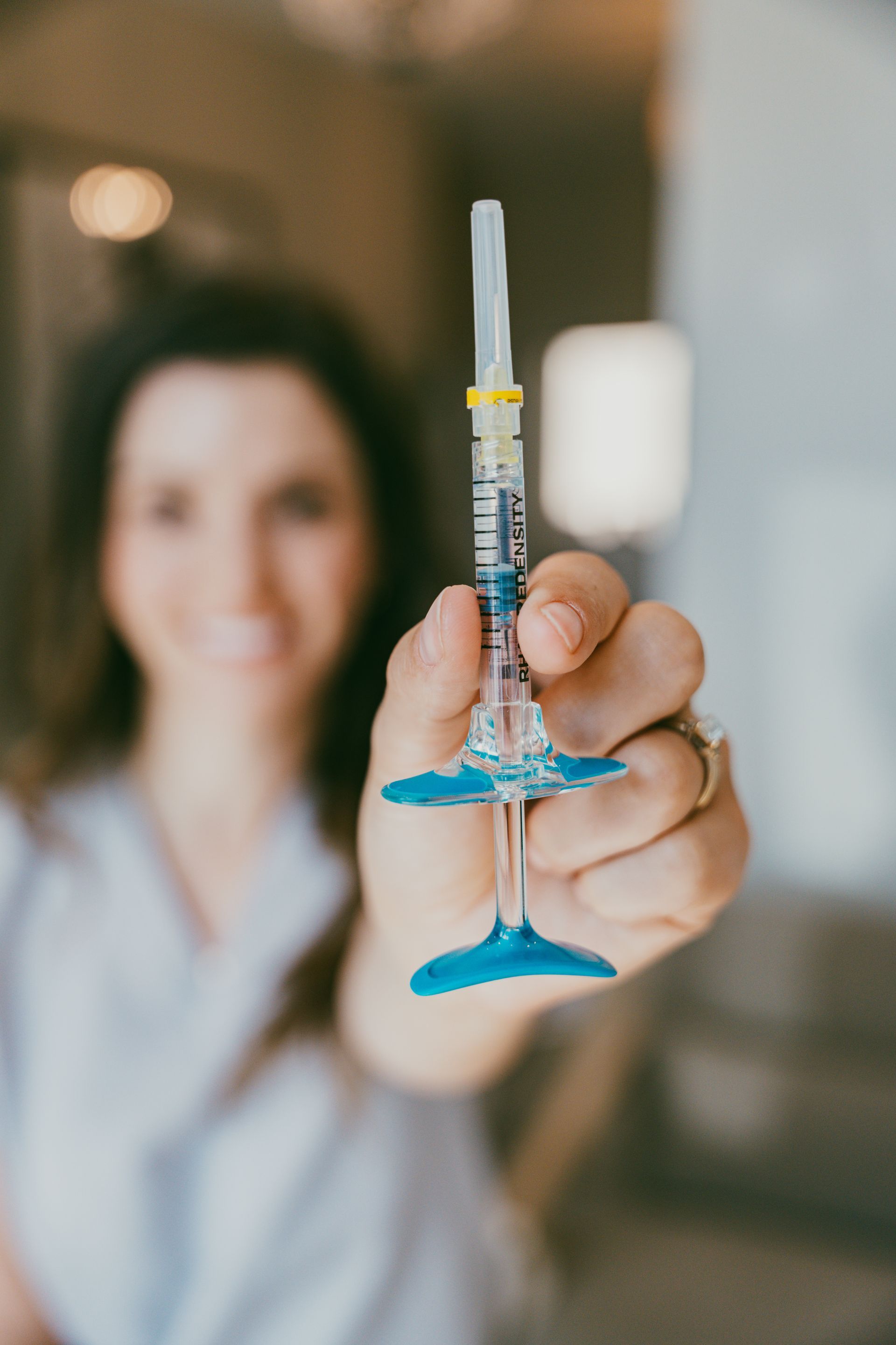Hand holding a syringe with blue cap, with a blurred person in a lab coat in the background