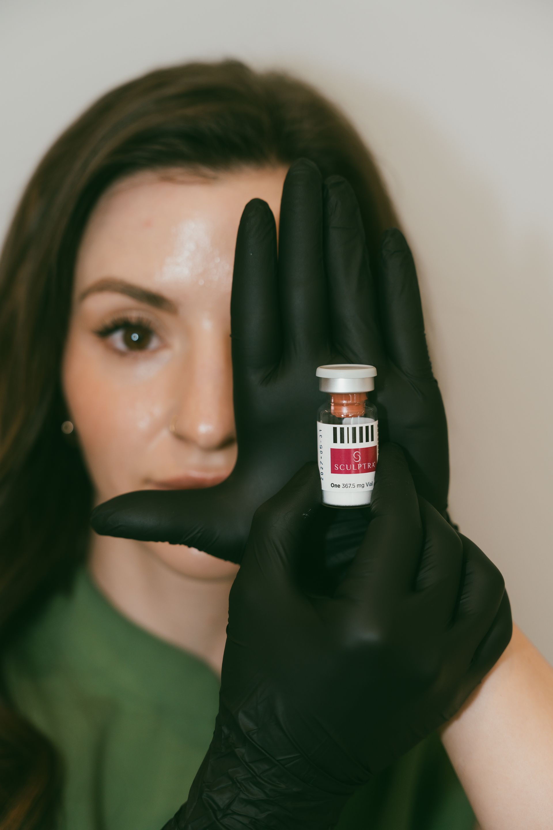 Woman holding up a gloved hand with a small vial in front of her face against a plain background