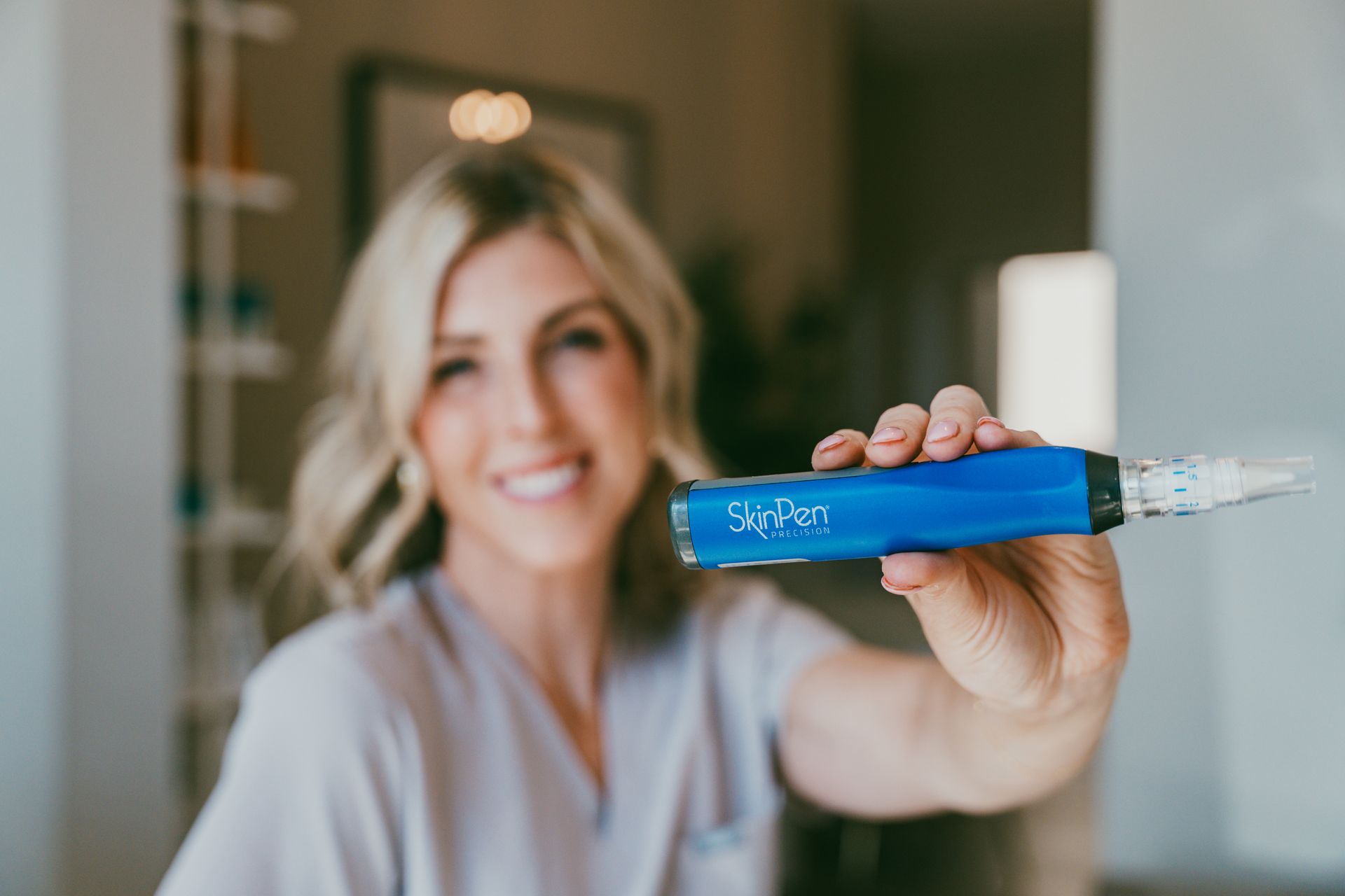 Woman holding a blue vape pen toward the camera indoors, smiling in soft focus.