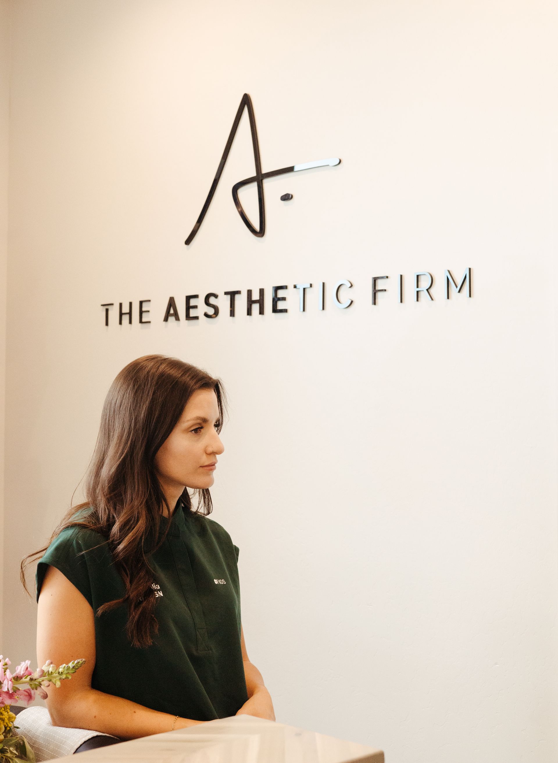 Woman seated beside wall logo reading “The Aesthetic Firm” in a bright office.