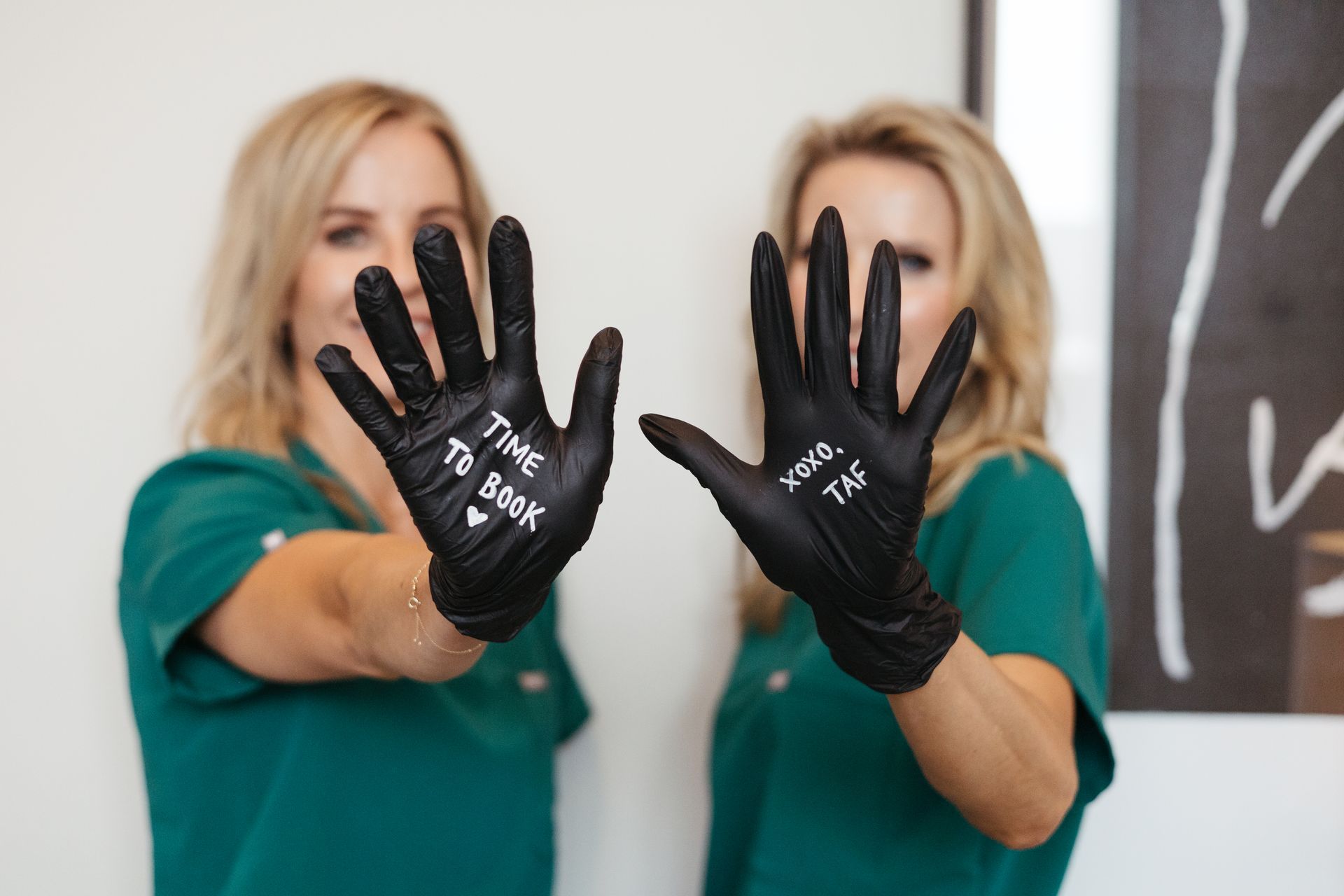 Two women in teal shirts hold up black gloves toward the camera in a bright room