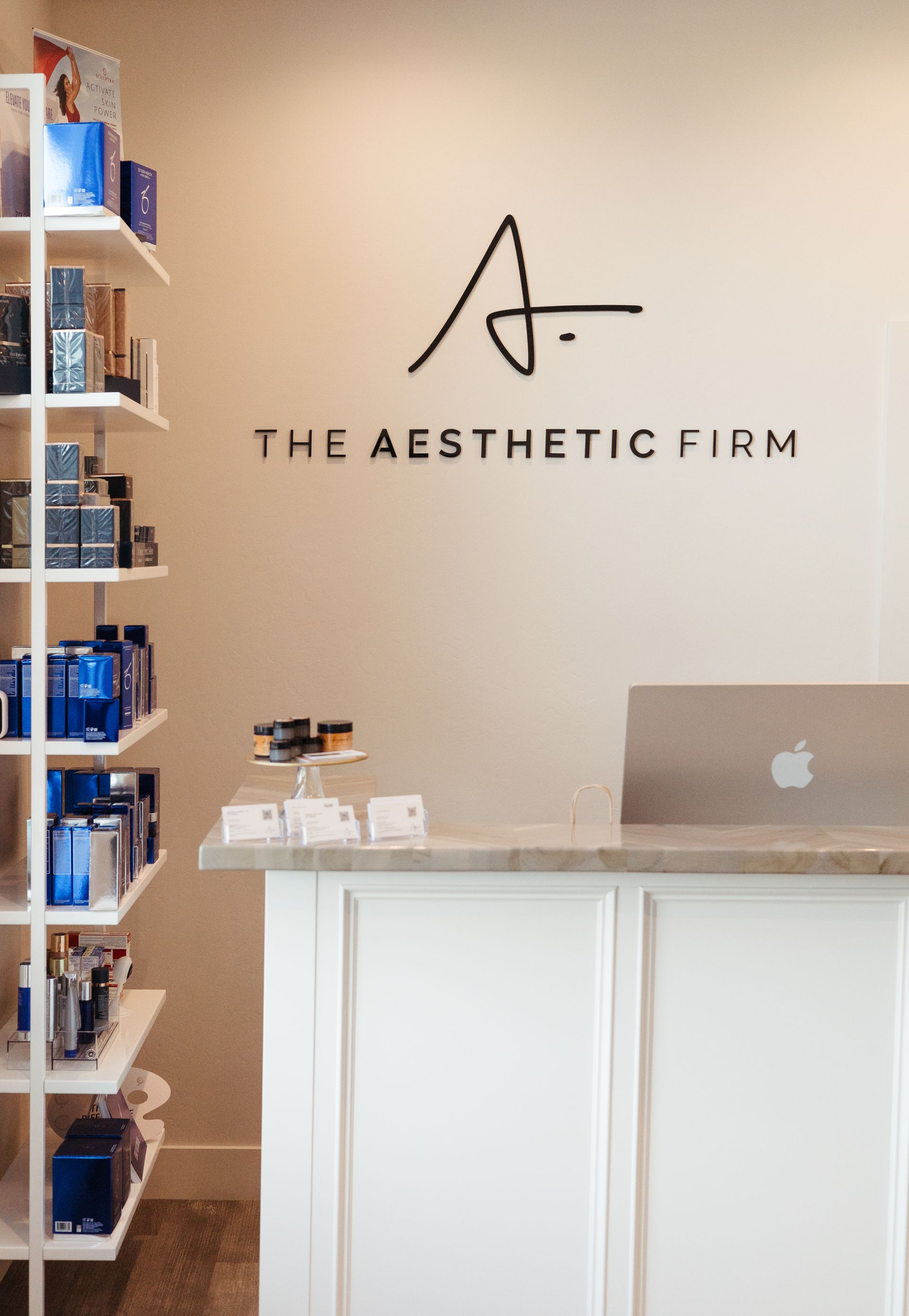 Reception desk at The Aesthetic Firm with white counter, laptop, and shelving display.