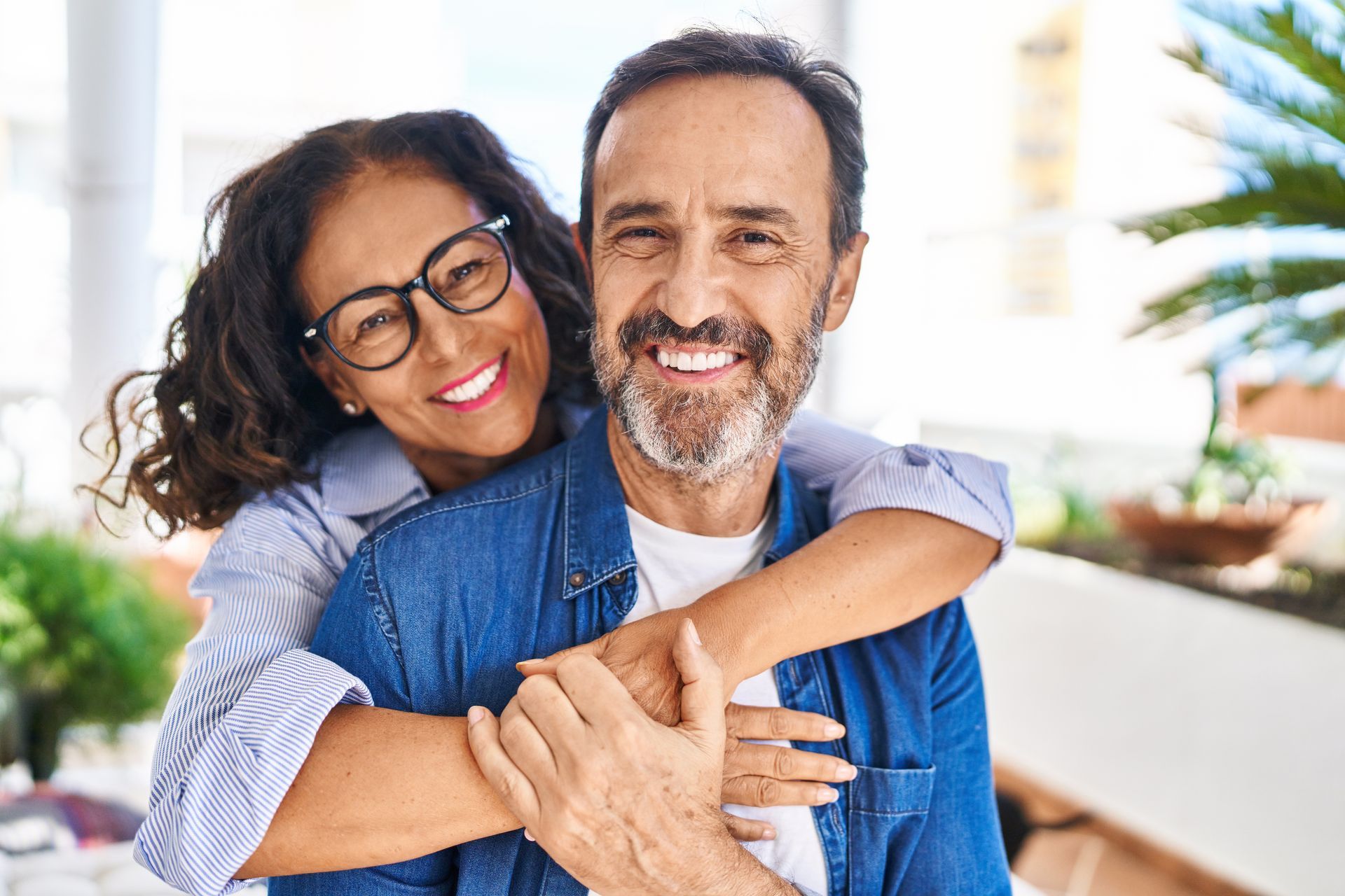 Smiling couple hugging indoors, with the man in a denim shirt and the woman wearing glasses.