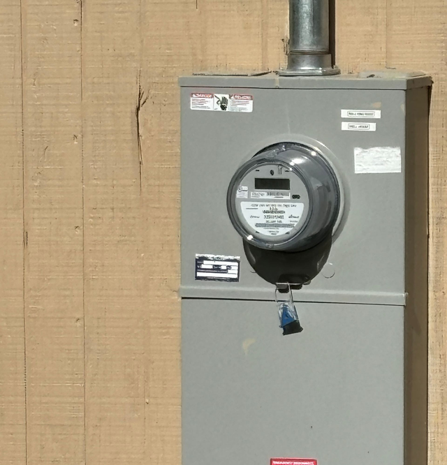 Gray electric meter box mounted on a tan wall with conduit above it
