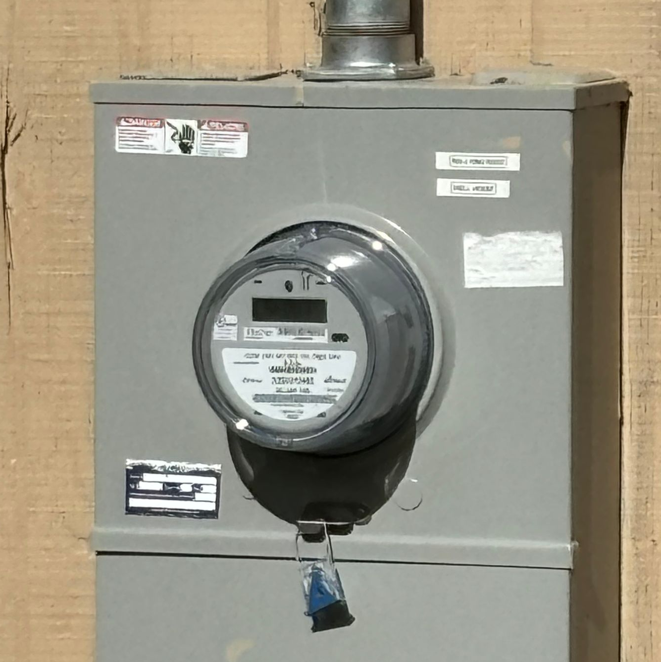 Gray electric meter box with round glass meter and warning labels, mounted on a wall
