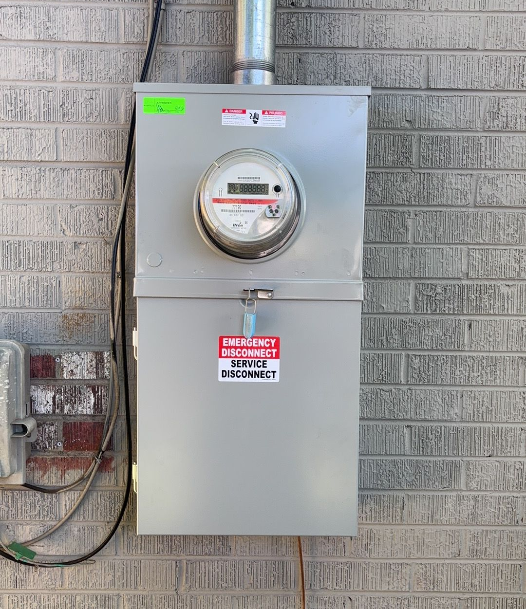 Gray electrical meter box mounted on a brick wall with conduit and warning labels