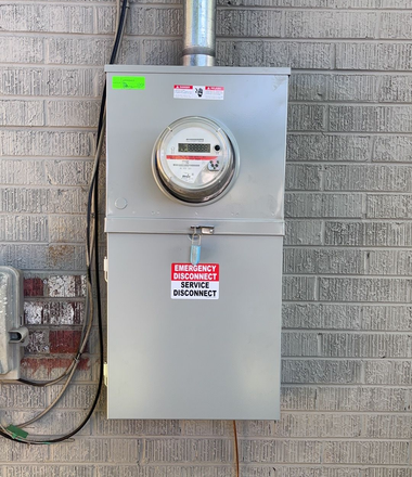 Gray electrical meter box mounted on a brick wall with conduit and warning labels