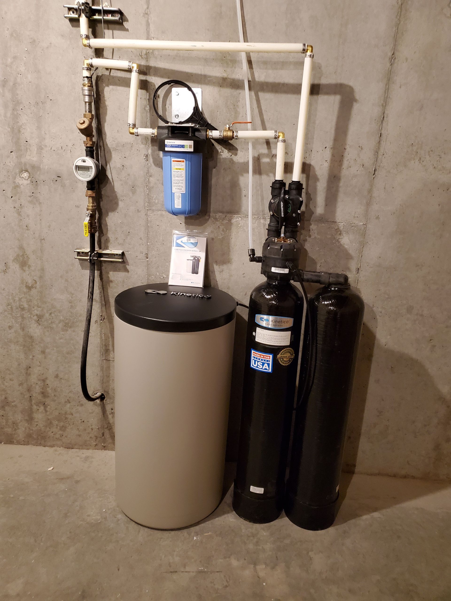 Water filtration system in a basement. Beige tank, black tanks, blue filter, white pipes.