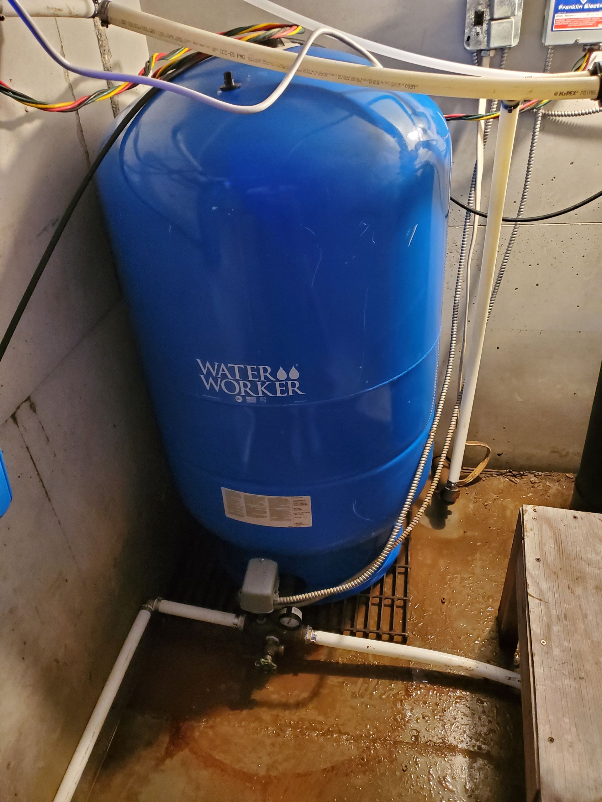 Blue water pressure tank in a basement setting, with white PVC pipes and a metal grate.