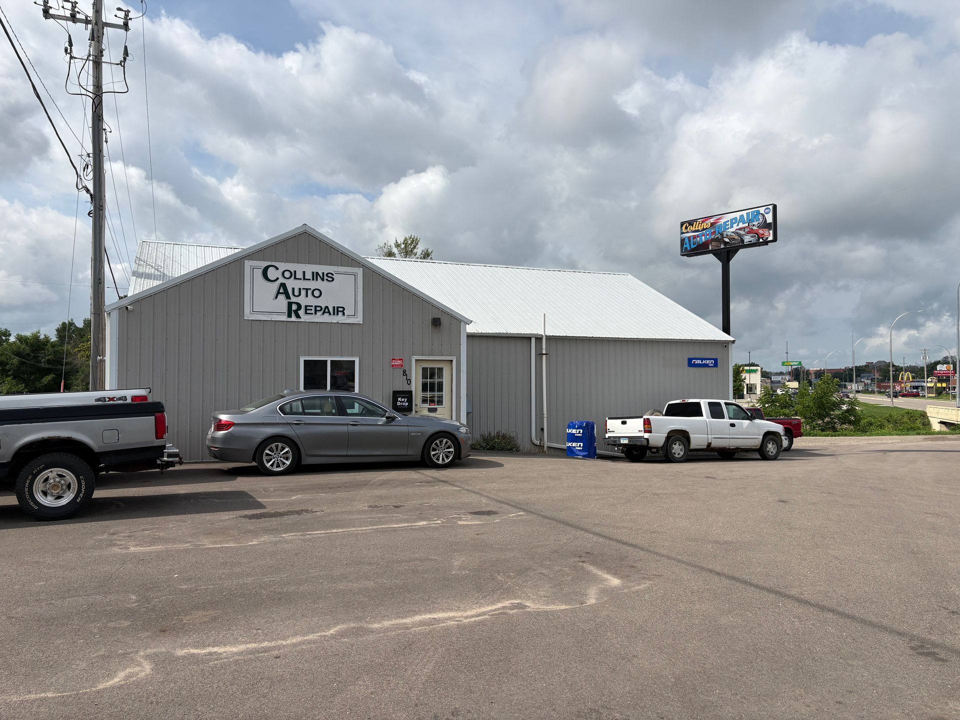 Single-story metal building with parked trucks and cars under a cloudy sky, with a roadside sign.