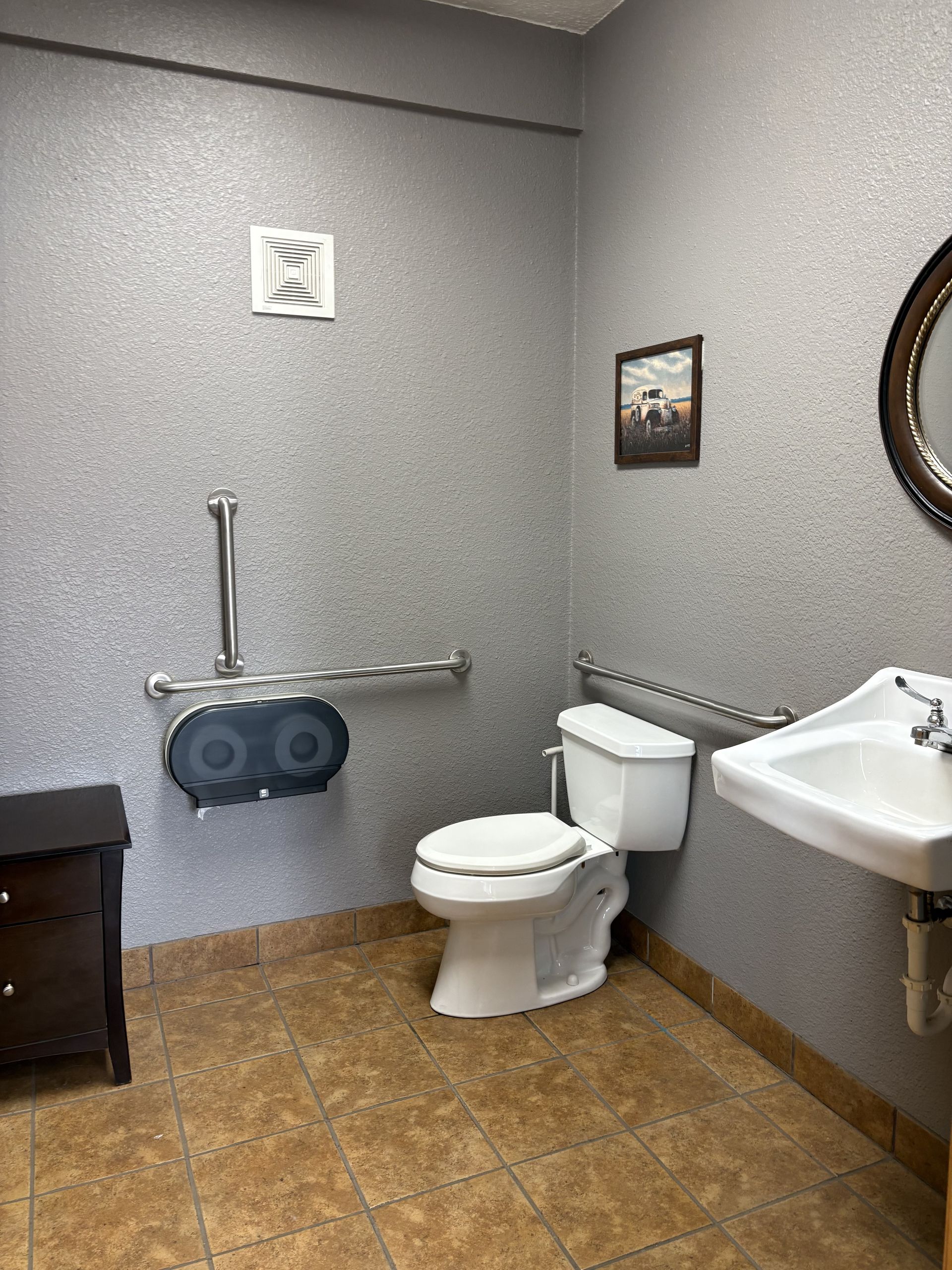 Accessible bathroom with toilet, wall-mounted sink, grab bars, and tan tile floor