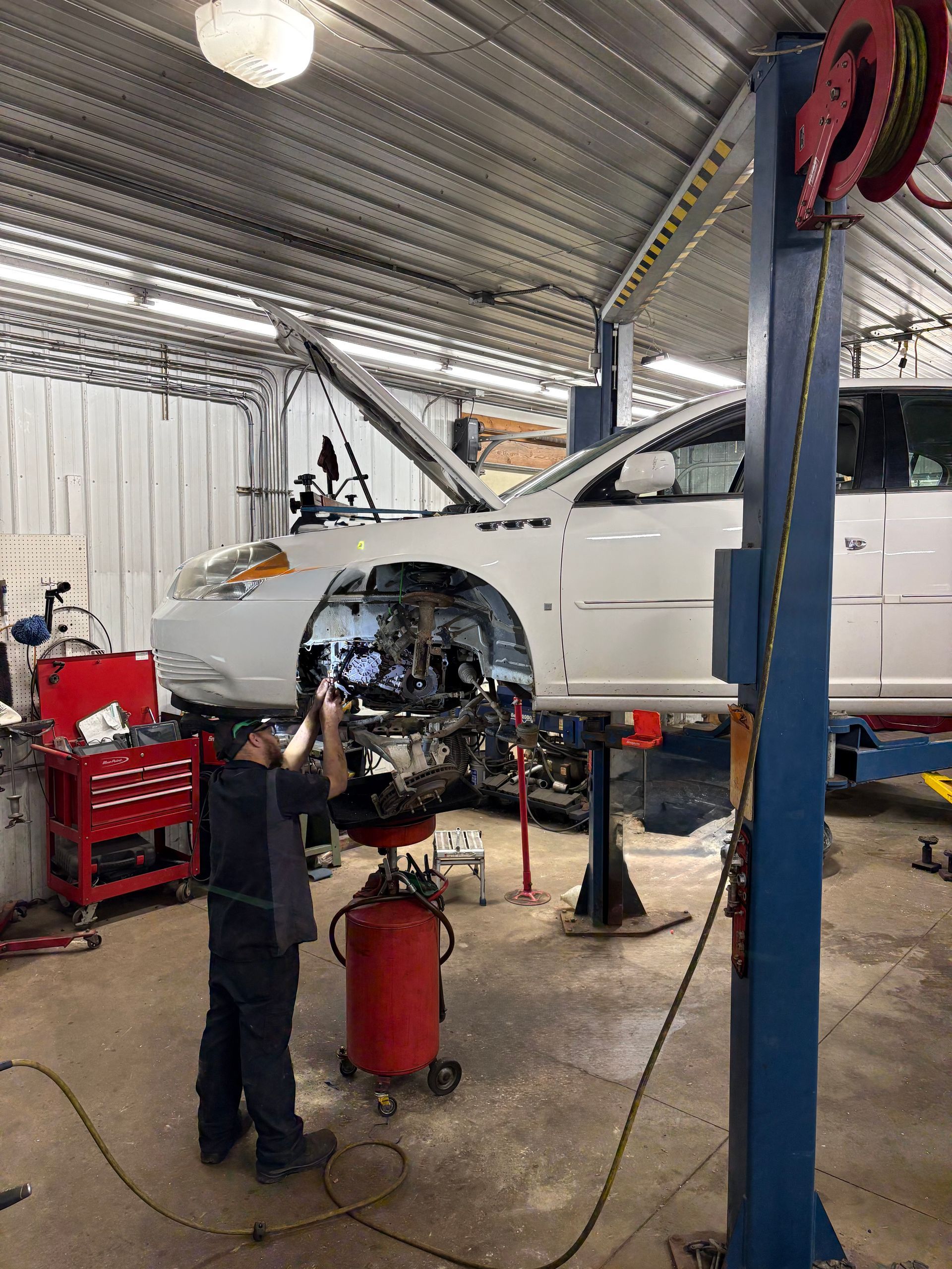 Mechanic working on a white car in a garage, with the front bumper removed and tools nearby.